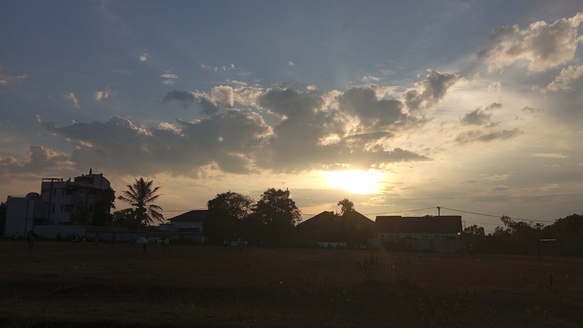 The image depicts a sunset scene in Uteunreutoh, Indonesia. The sky is filled with dramatic clouds, with the sun setting on the right side of the frame, casting a warm glow. In the foreground, a field of dry grass is visible, with several silhouetted figures scattered across it, seemingly engaged in playing soccer. To the left, a multi-story building stands out, with a palm tree nearby. Several other buildings and trees are visible in the middle ground and background, all silhouetted against the vibrant sky. The overall atmosphere is serene, with the fading daylight creating a peaceful ambiance.