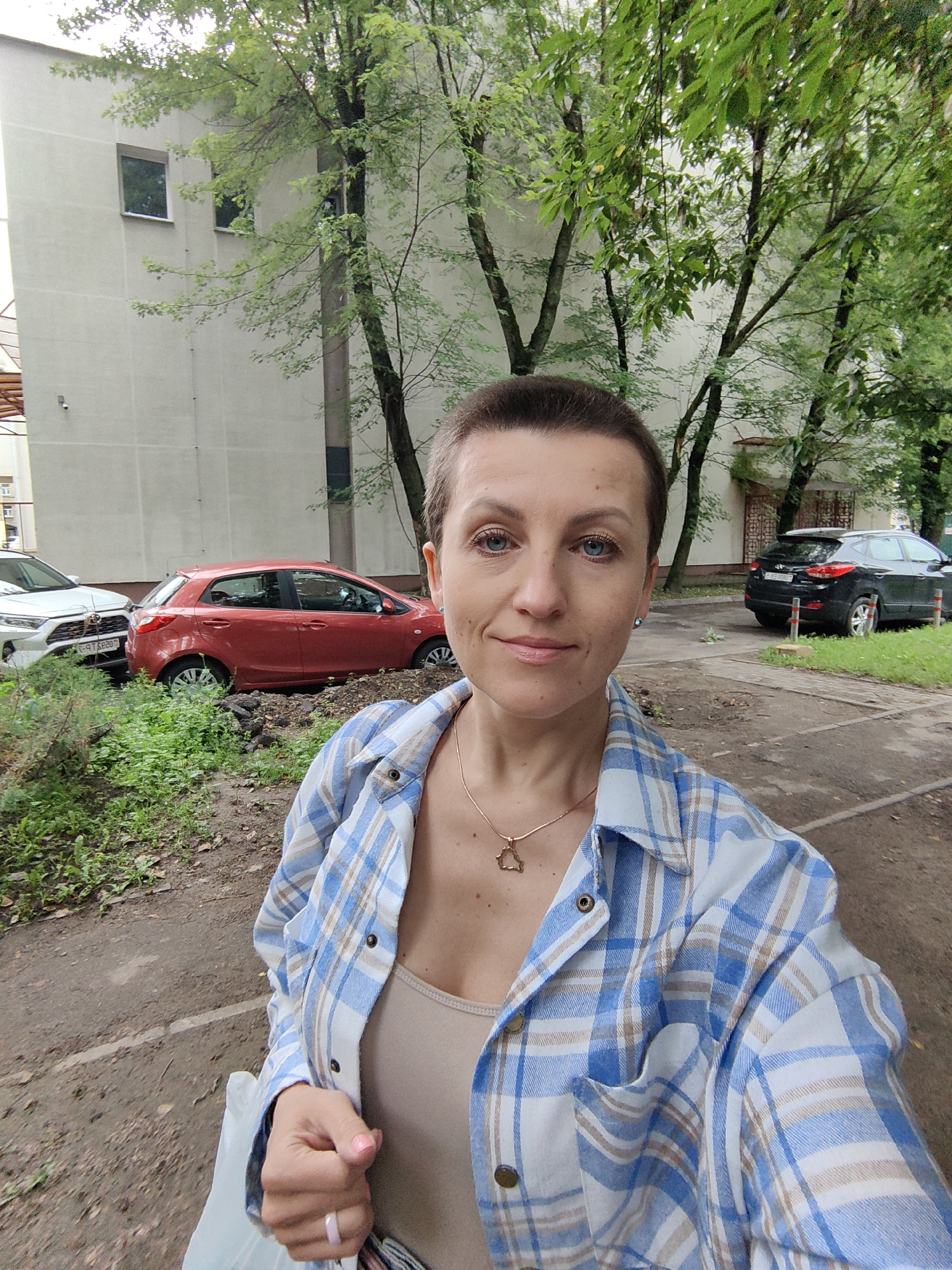 A woman with short brown hair and blue eyes is taking a selfie outdoors in front of a building. She is wearing a blue and white plaid shirt over a tan top and a gold necklace with a small pendant. Her left hand is holding a white bag, and she is wearing a pink-toned ring on her left ring finger. In the background, there is a light-colored apartment building, trees with green foliage, and several parked cars, including a red hatchback and a black SUV. The scene appears to be a residential area. The image has a timestamp that indicates it was taken at 2023-08-07 13:06:24.
