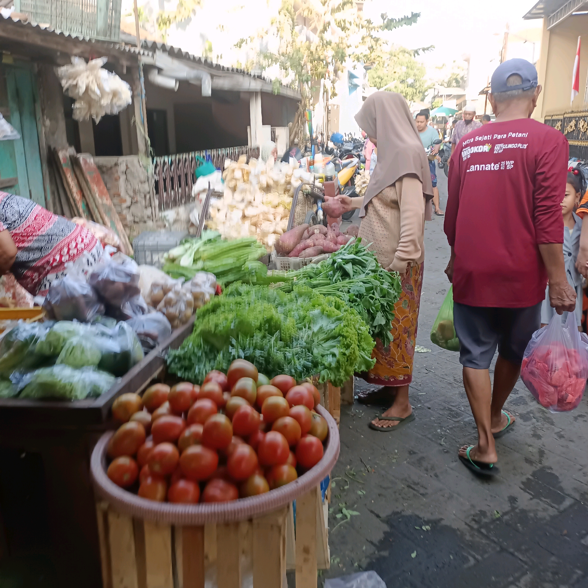 A street market scene in Surabaya, Indonesia, features stalls filled with fresh produce. In the foreground, a wooden crate overflows with ripe red tomatoes. To the left, a person wearing a patterned top and a headscarf is partially visible behind a display of bagged leafy greens and other vegetables. Further back, a woman in a light brown hijab and patterned skirt examines sweet potatoes at a stall. Behind her, a man wearing a maroon long-sleeved shirt and grey shorts carries two plastic bags, one containing green items and the other red meat. Other shoppers and market stalls are visible in the background, along with parked motorcycles, suggesting a bustling commercial area. The ground is wet and appears to be paved with dark asphalt or concrete. Sunlight filters through trees overhead, illuminating the scene.