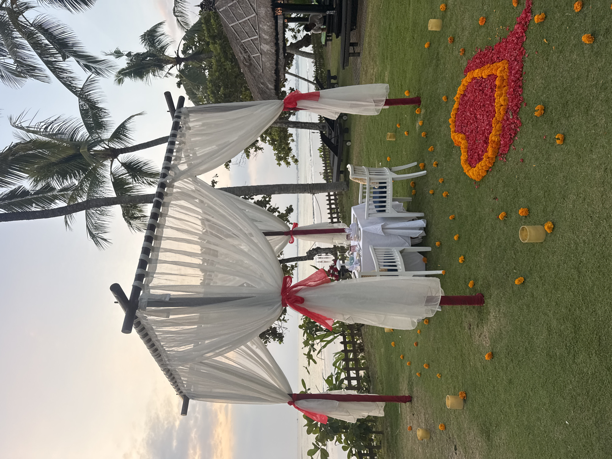 An outdoor romantic dining setup is depicted on a grassy lawn in Kuta Selatan, Indonesia, with a beach and ocean visible in the background. The scene is illuminated by the soft light of an overcast sunset. A white canopy, draped with sheer white fabric and adorned with red ribbons, is set up over a white-clothed dining table. The table is set for two with white plates, cutlery, and glasses. White chairs are placed around the table, and a bouquet of red flowers sits on the table.
The lawn is scattered with small orange flowers and several yellow cylindrical candle holders, some with candles lit. In front of the dining setup, a large heart shape is formed on the grass using red flower petals and bordered by orange marigolds. Beyond the lawn, a wooden fence separates the area from a strip of green foliage, with lounge chairs and a thatched-roof gazebo visible further back. The ocean waves are gently rolling onto the shore, and the sky is a pale gradient of orange and blue. A few palm trees are present, adding to the tropical ambiance. One person is visible in the background, near the gazebo.