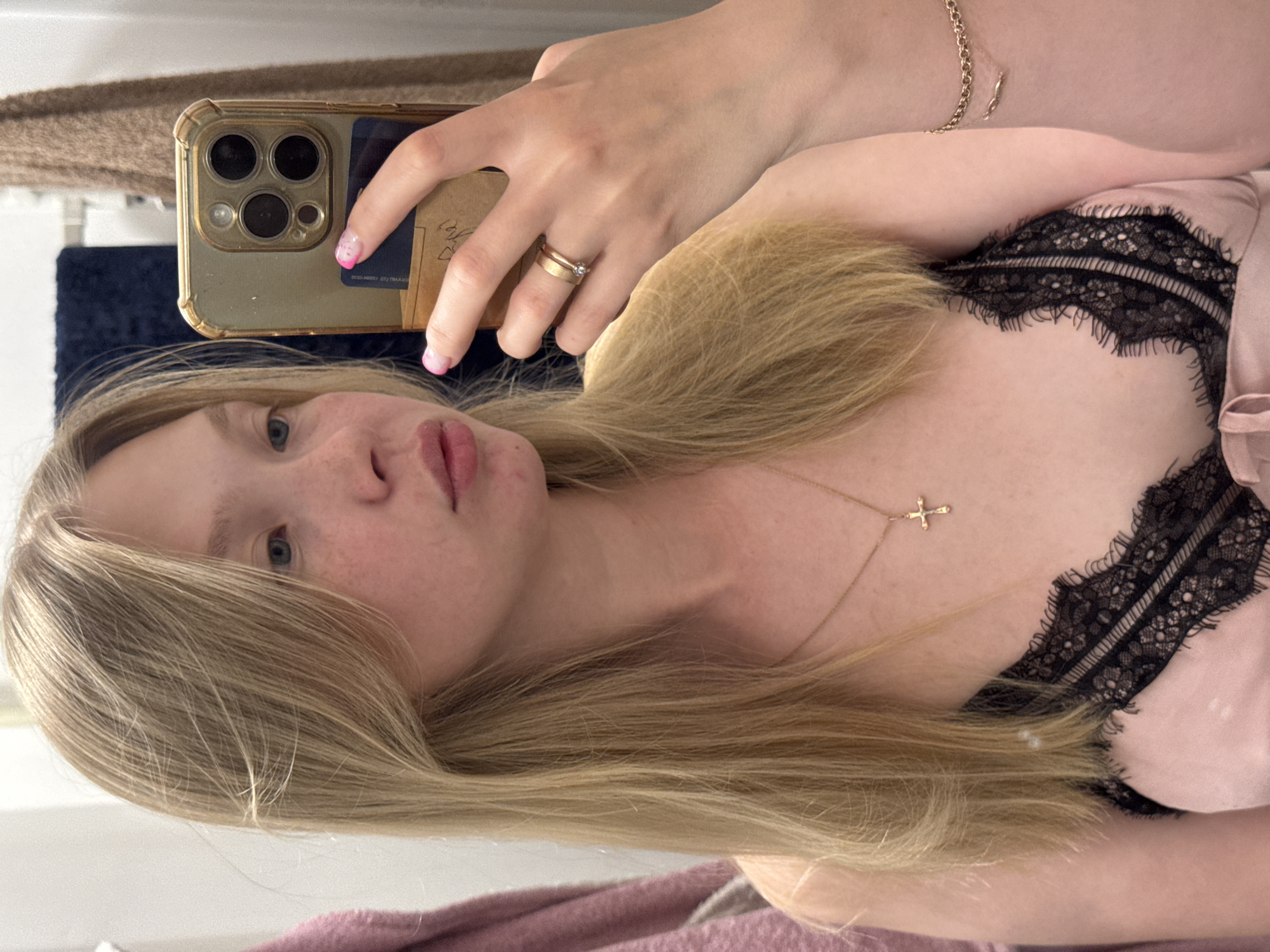 This is a vertically oriented, close-up selfie taken by a young woman. She has fair skin with freckles, light blue eyes, and long blonde hair. She is wearing a pale pink silk camisole with black lace trim and a delicate gold necklace with a small cross pendant. She is also wearing two gold rings on her left hand, one with a small diamond. Her fingernails are painted pink with white tips.
She is holding a smartphone in her right hand, and the phone's camera is capturing her reflection. The phone has a gold case and a prominent camera array. Behind her, a blurred background suggests an indoor setting, possibly a bedroom or dressing area, with hints of fabrics and what appears to be a mirror. The lighting appears soft, possibly natural light from a window, suggesting it could be daytime. There is no discernible text in the image. The woman's expression is neutral, and she seems to be posing for the photo. The overall impression is casual and personal.