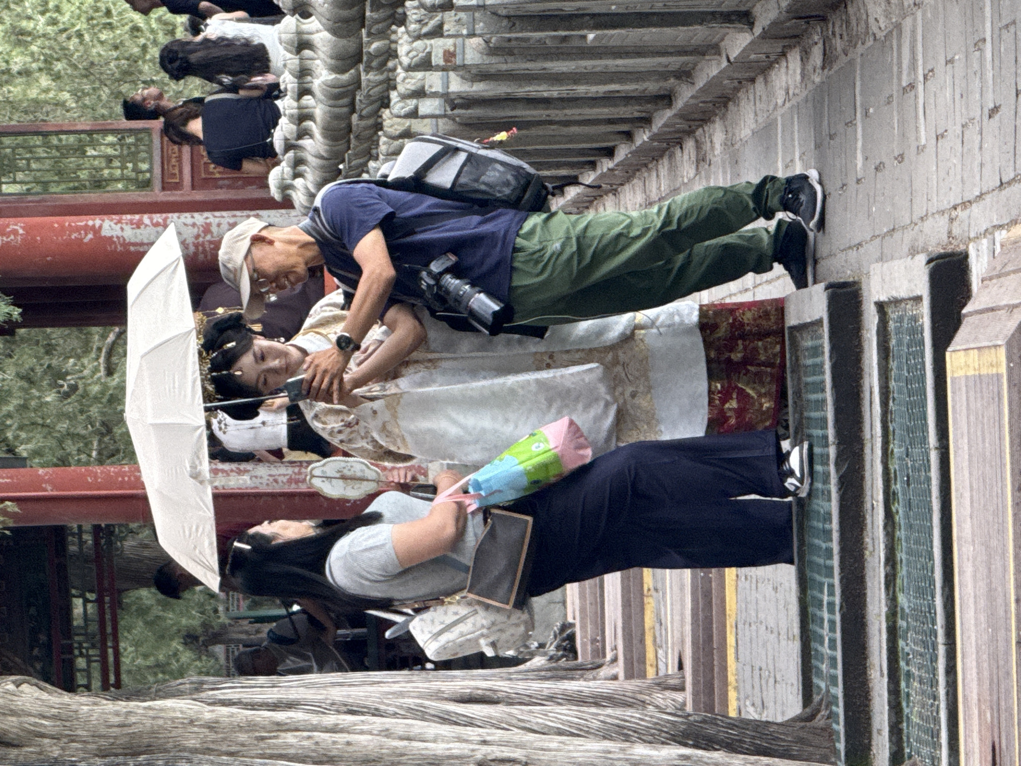 This image captures a daytime scene on a paved walkway within the Summer Palace (Yiheyuan) in Haidian District, China. In the foreground, a young adult female in elaborate traditional Chinese attire, including a white robe with gold embroidery and an ornate dark hairstyle, holds a smartphone, looking at its screen. A white umbrella is held above her head for shade. To her right, a middle-aged male, wearing a beige baseball cap, glasses, a dark blue short-sleeved shirt, olive green pants, and a grey backpack, leans in to view the smartphone, his right hand near hers. A DSLR camera with a long lens hangs from a strap around his neck.
To the left of the central figures, an adult female with dark hair, wearing a light grey ribbed short-sleeved top and dark blue wide-leg pants, stands with a light-colored patterned backpack on her left shoulder, a dark brown cross-body bag, and a multicolored (pink, light blue, green) tote bag in her left hand. She is looking towards the central group. Her footwear consists of white sneakers with black accents. The man with the camera wears black athletic shoes.
The immediate environment includes a prominent tree trunk with textured bark on the far left, dark green metal drainage grates embedded in the walkway, and a grey ornate stone balustrade with numerous vertical pillars extending along the right side. In the background, traditional Chinese architecture with red pillars and dark, intricate wooden lattice work is visible amidst dense green foliage. Several other individuals, partially obscured and blurred, are present in the background, indicative of a public tourist location. The lighting is bright daylight, casting visible shadows.