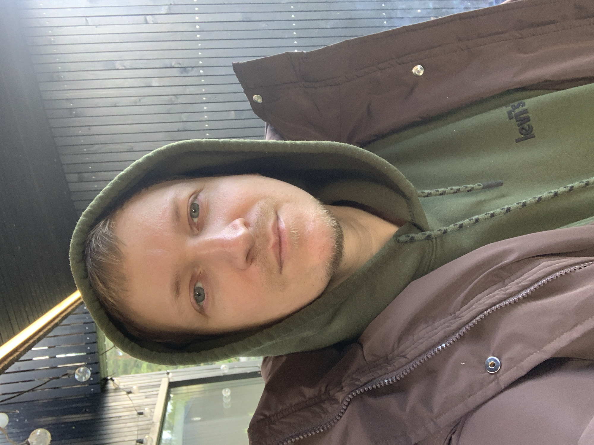 The image shows a man wearing a brown jacket over a green hooded sweatshirt. He has short, light brown hair and a light beard. The background consists of a dark wooden wall and a ceiling with string lights. The man is looking directly at the camera. Based on the provided information, this scene is located in the village of Myazhantsy, within the Mezhan Rural Council, in Belarus.