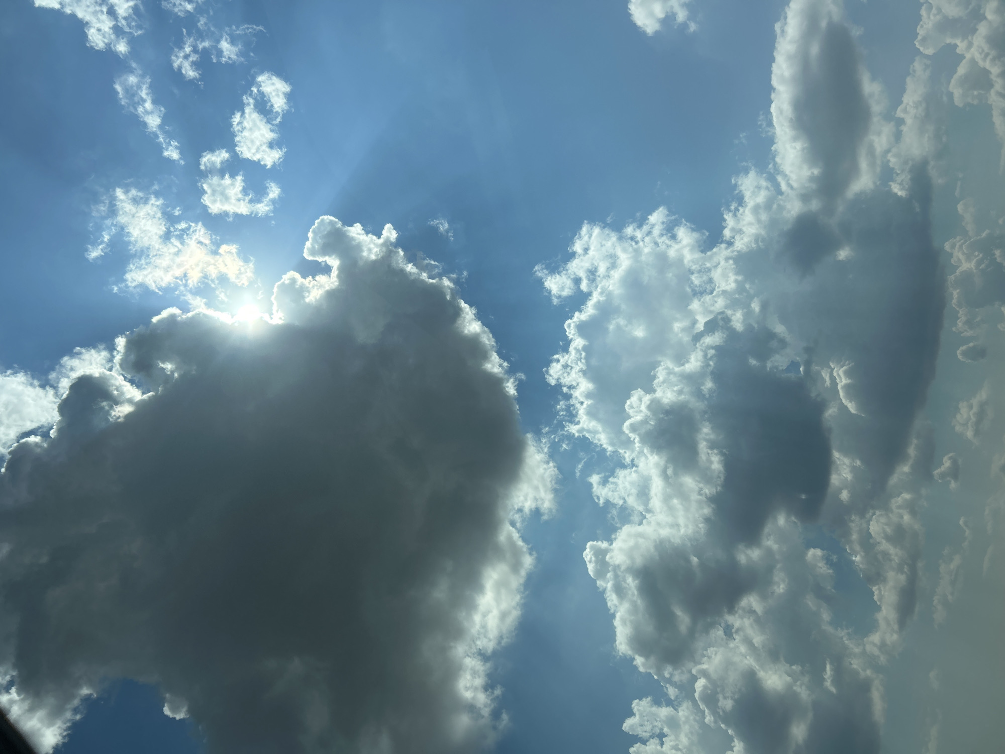A photograph captures a bright blue sky filled with dramatic clouds. The sun is partially obscured by a large, dark cloud on the left side of the frame, casting rays of light that fan out across the sky. Smaller, white, fluffy clouds are scattered throughout the scene, some appearing more distant and wispy. The overall impression is one of a dynamic and somewhat turbulent sky, likely taken on a day with changing weather conditions. The image does not contain any discernible urban features or people, and therefore cannot be localized to Загорци, Bulgaria, or any other specific urban location.