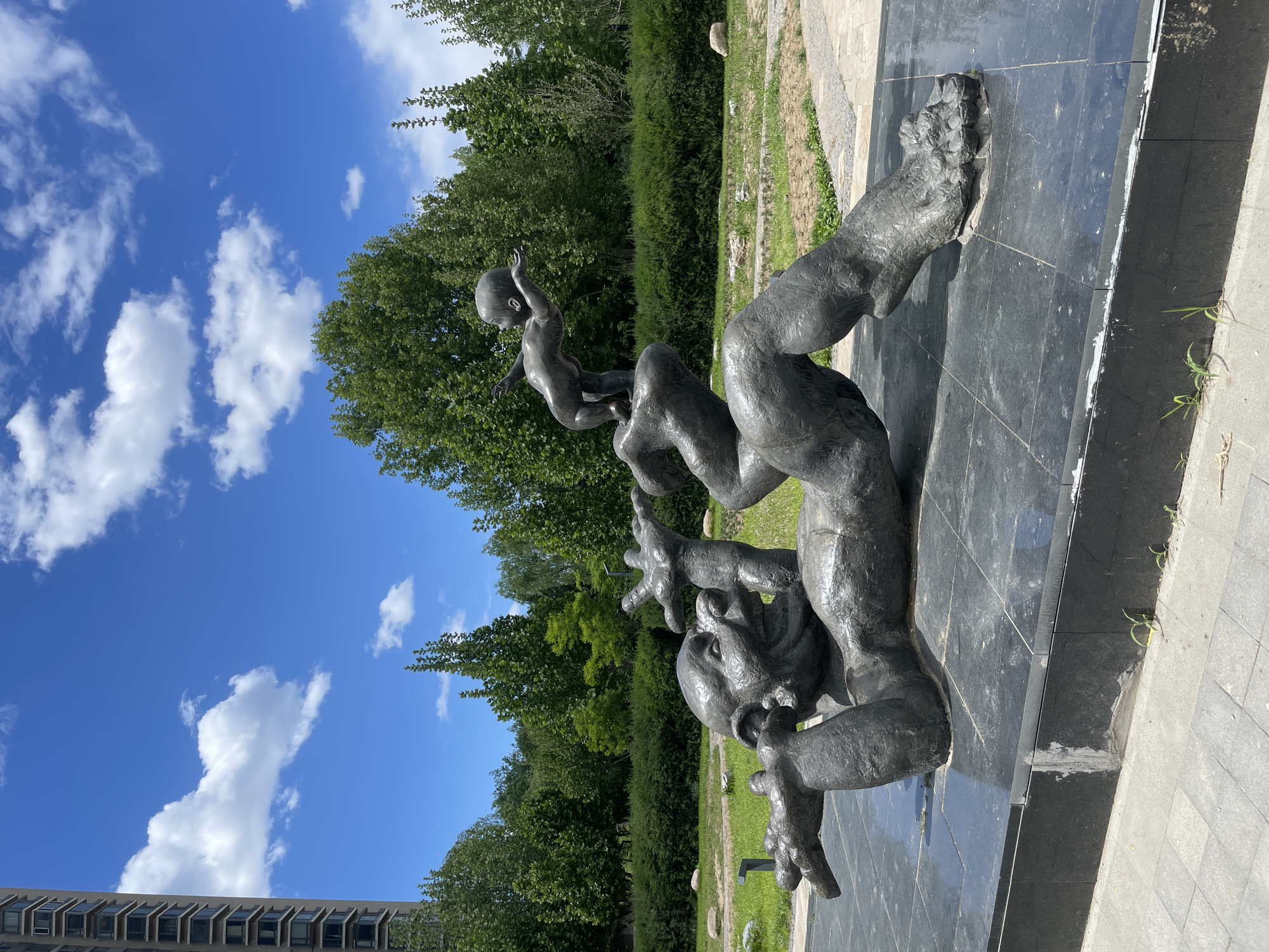 The image shows a bronze sculpture of a man supporting a child on his shoulders, located outdoors. The sculpture is on a black tiled platform. The sky is blue with scattered white clouds. A line of trees and green vegetation is visible in the background, along with a portion of a building on the left side. The location is Changping District, China, based on known landmark elements.