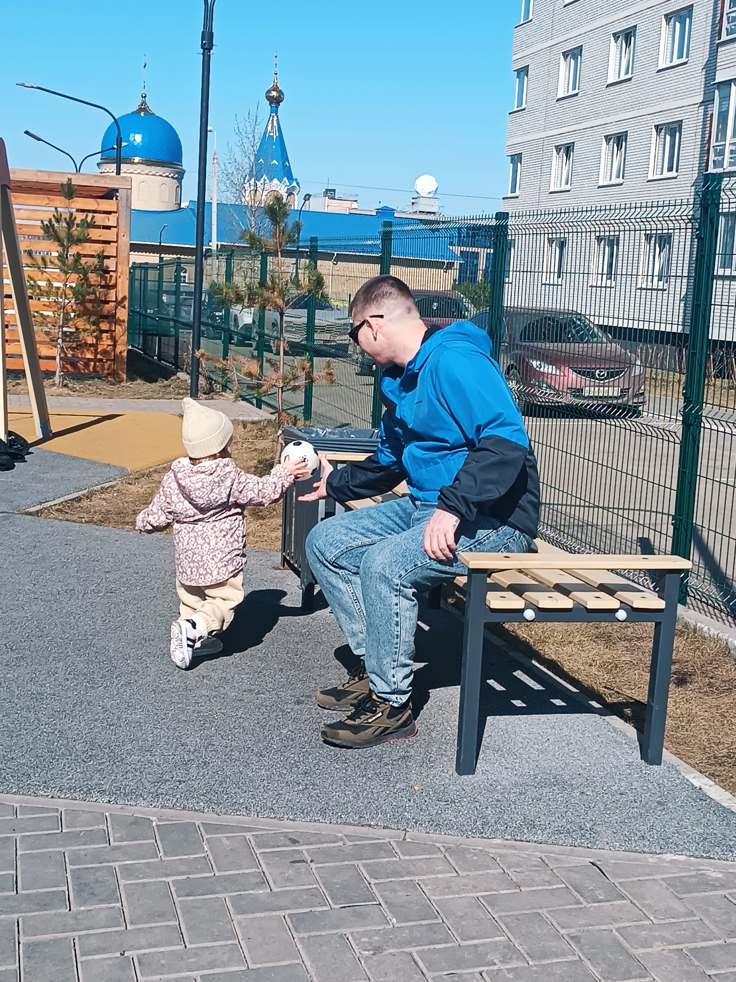 A toddler walks towards a seated man, extending their arms to hand him a black and white soccer ball. The toddler wears a light beige hat, floral-patterned jacket, and matching pants. The man is seated on a gray and wood park bench; he is wearing jeans, brown shoes, and a blue jacket with black sleeves. The photograph is taken outdoors on a sunny day, with the sky a clear, light blue.
The environment includes buildings, fences, and landscaping typical of an urban setting. In the background, there is a chain-link fence, apartment buildings with light-colored facades and dark windows, and landscaping consisting of grass and small trees. In the distance, there are two buildings with blue domes. According to metadata, this image was taken in Izhevsk, Russia.
