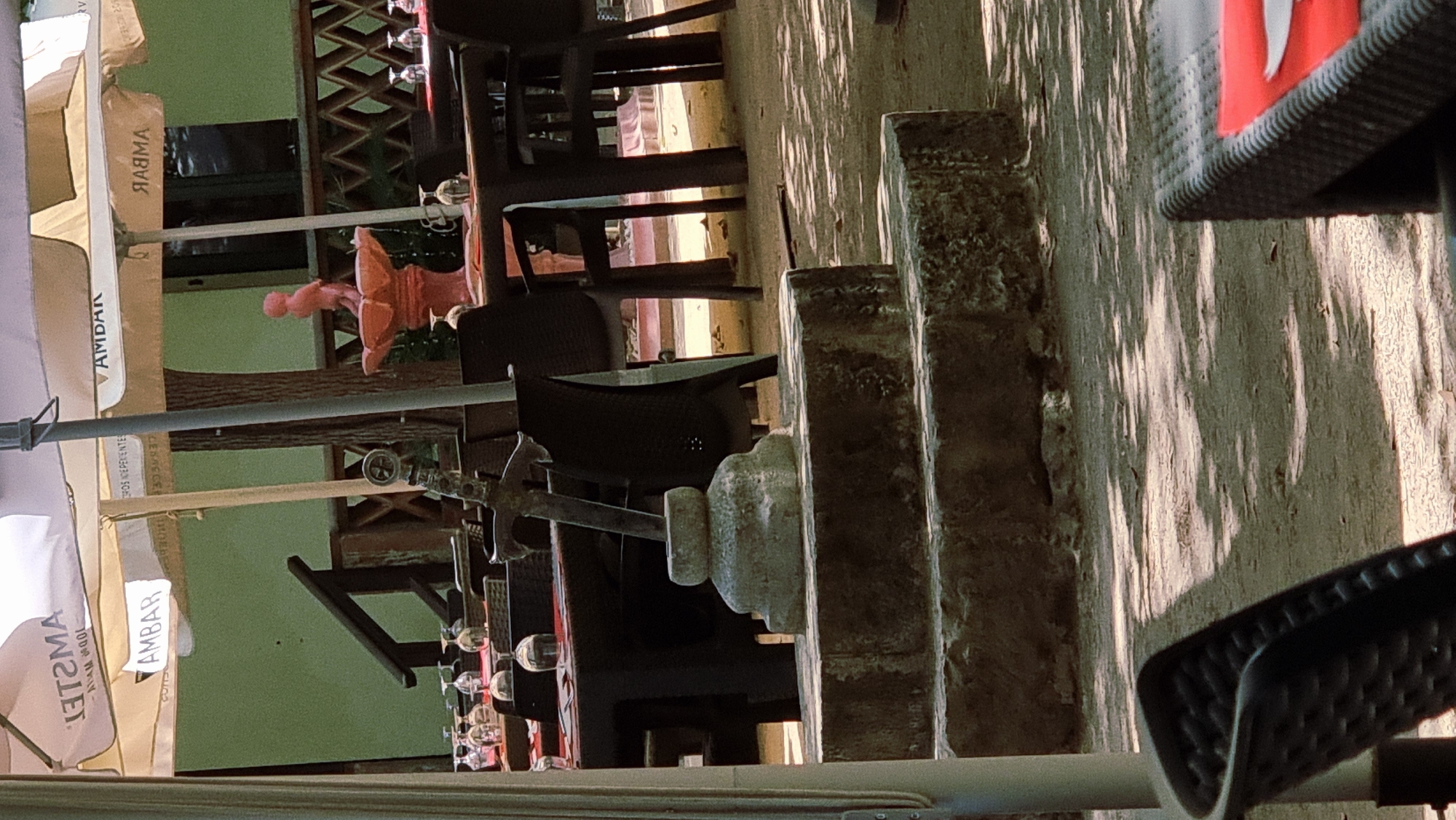 The image shows an outdoor seating area of a restaurant or cafe. In the foreground, a stone pedestal with a metal structure resembling a sword hilt is visible. The area is furnished with tables and chairs, some of which are covered by umbrellas. The umbrellas bear the logo "AMBAR". In the background, there is a pink statue of a child on a fountain. The ground appears to be made of sand or gravel, with scattered leaves. The scene is bathed in sunlight, creating shadows on the ground. The overall impression is that of a relaxed, outdoor dining environment. Given the context of the "AMBAR" branding, this location is likely in Spain. While this specific image does not contain elements that definitively place it in Besalú, Spain, such restaurants are common in historic towns like Besalú.