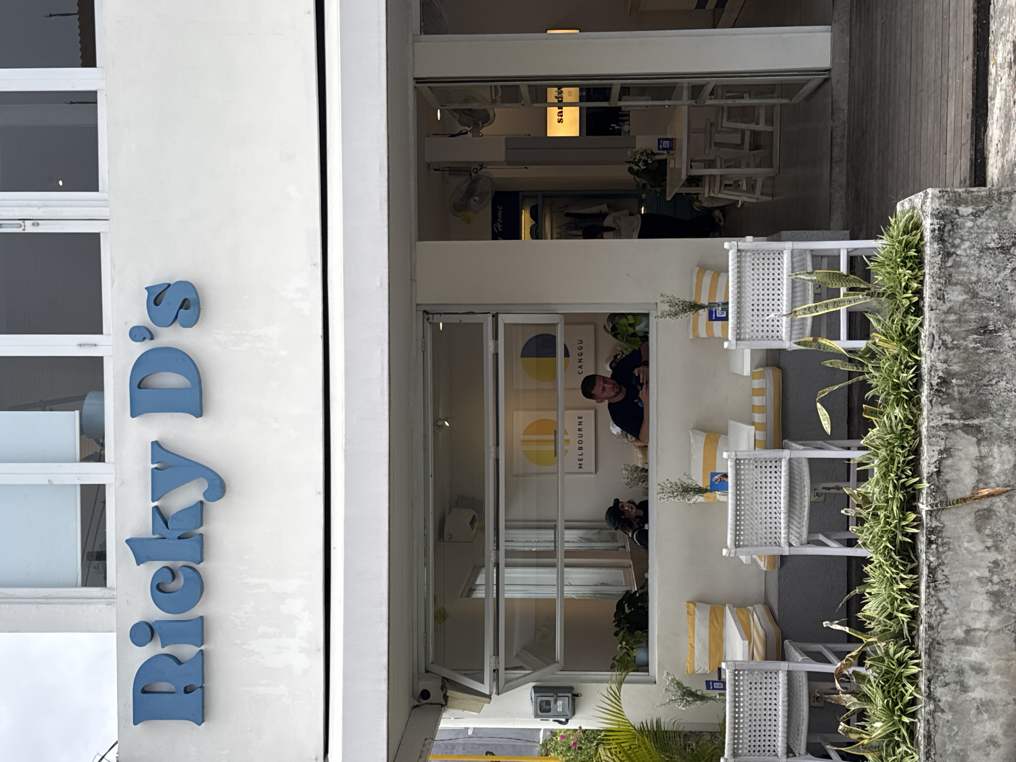 The image displays the bright, white-walled facade of a cafe named "Ricky D's" in what appears to be a daytime setting in North Kuta, Indonesia. The building features large windows, showcasing an inviting interior.
The main sign, "Ricky D's," is prominently displayed in a stylized blue font on the white exterior wall. An outdoor seating area is visible on the right side of the entrance (after rotation), furnished with two white woven chairs, each adorned with yellow and white striped cushions. A concrete planter box filled with lush green foliage separates this seating area from an unseen pathway or street.
Through the large windows, the cafe's interior is discernible. More white tables and chairs are arranged inside, suggesting ample seating. Two ceiling fans are mounted overhead. A rectangular illuminated sign inside displays the text "SANDI" (or "SANTI") and a QR code. Below this, closer to the outdoor seating, another framed artwork or sign is visible, featuring a circular design with yellow and blue/grey segments, and the words "CANGGU" and "MELBOURNE," providing strong location and perhaps origin cues.
Several people are within the cafe. One man with dark hair and a beard, wearing a dark t-shirt, is clearly visible leaning out from one of the lower windows, looking towards the outside. Another person is partially visible deeper inside the cafe. The scene suggests a relaxed atmosphere at a local establishment, likely catering to customers in the Canggu area.