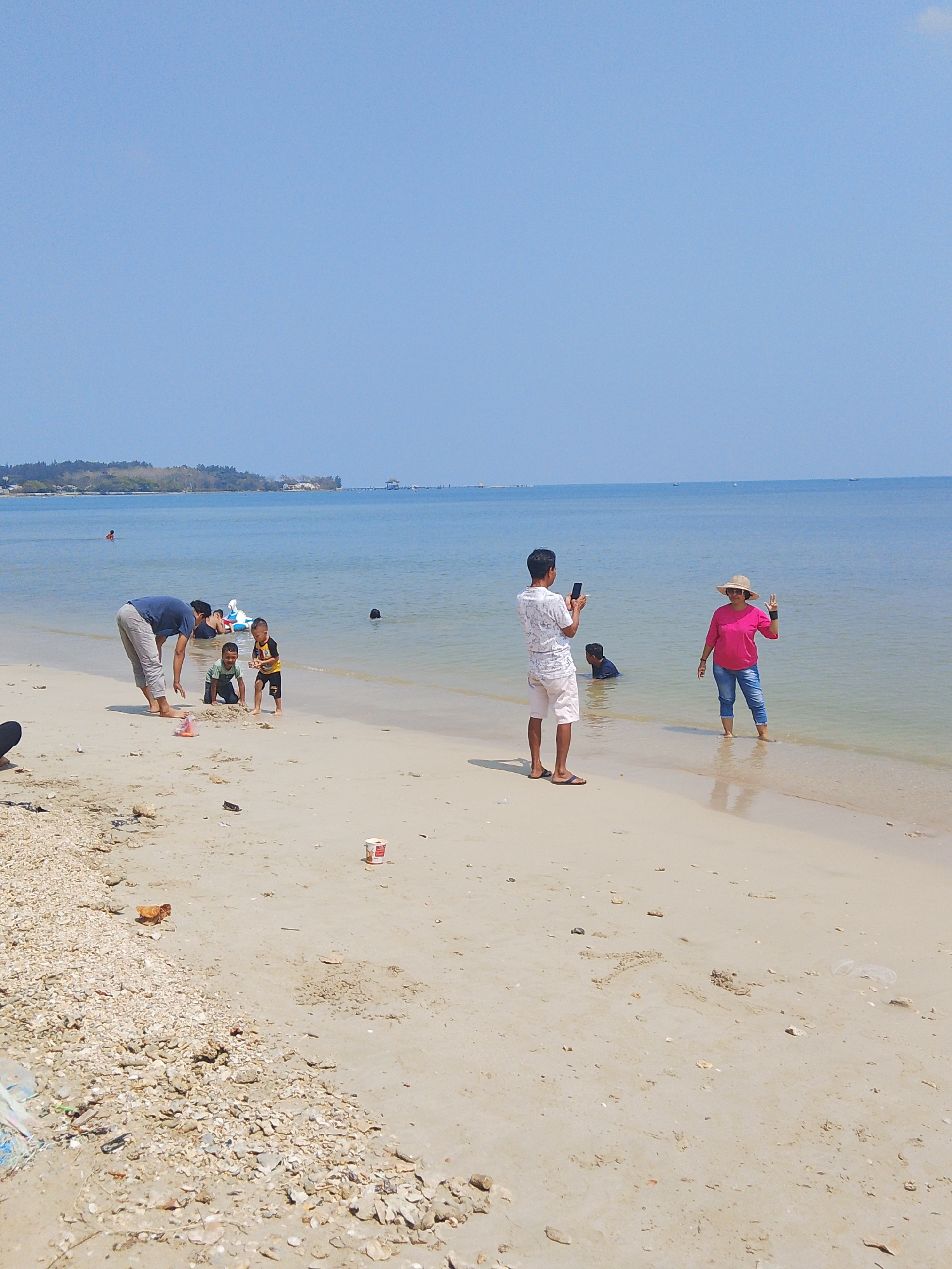 The image depicts a beach scene in Pejagan, Indonesia. The water is a clear blue, meeting a sandy beach scattered with shells and debris. Several people are present. In the foreground, a man in a dark blue t-shirt and grey pants bends down near two young boys playing in the sand. One boy is wearing a green shirt, and the other a yellow and black shirt. In the middle ground, a man in a white printed t-shirt and light-colored shorts is holding up a phone, seemingly taking a picture. To his right, a woman in a bright pink t-shirt and blue jeans, wearing a straw hat, stands in the shallow water and makes a peace sign with her left hand. Another person is visible in the water behind her. Further out in the water, other individuals can be seen, some swimming or wading. In the background, a tree-covered coastline stretches across the horizon, with structures and a pier visible in the distance. The sky is a clear, pale blue.