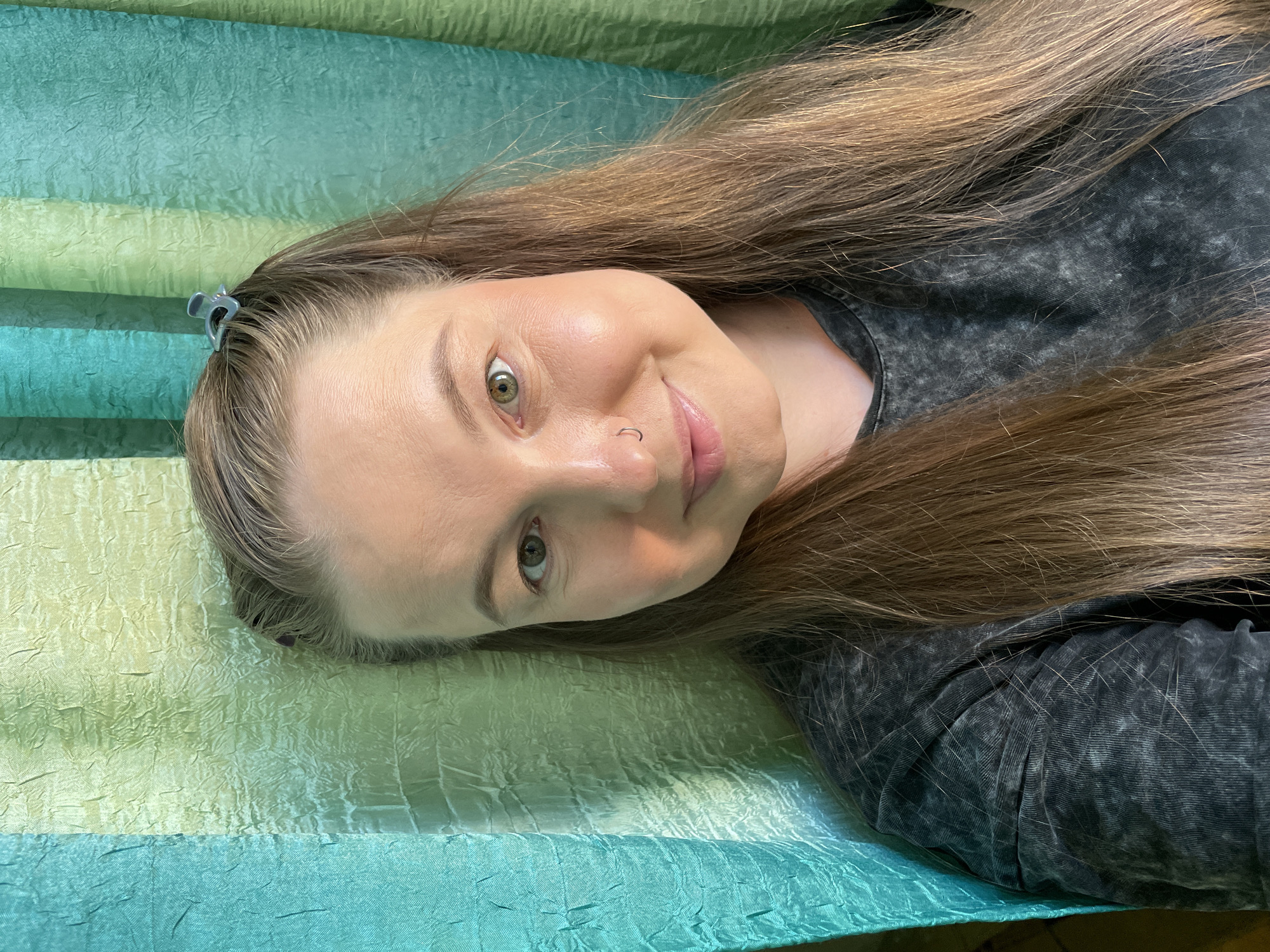 A young woman is depicted lying down, with her head positioned towards the left of the frame and her body extending to the right, looking directly at the camera. She has long, straight hair, appearing to be light brown with subtle highlights, which spreads out around her. Her eyes are hazel or green, and she sports a small, silver hoop nose ring on her left nostril. A small, grey, claw-like hair clip secures some of her hair on the top left side of her head.
The woman has a gentle, relaxed expression with a subtle smile. She is wearing a dark grey, short-sleeved top with a visibly textured or marled pattern.
The background is a vibrantly striped fabric surface, possibly a blanket or bedspread, featuring horizontal bands of crinkled texture. The colors transition from lighter lime-green tones to deeper teal and blue-green hues. The lighting in the image appears bright and even, consistent with an indoor setting during the day or under good artificial light. No text is visible in the media file. The provided location context is Izhevsk, Russia.