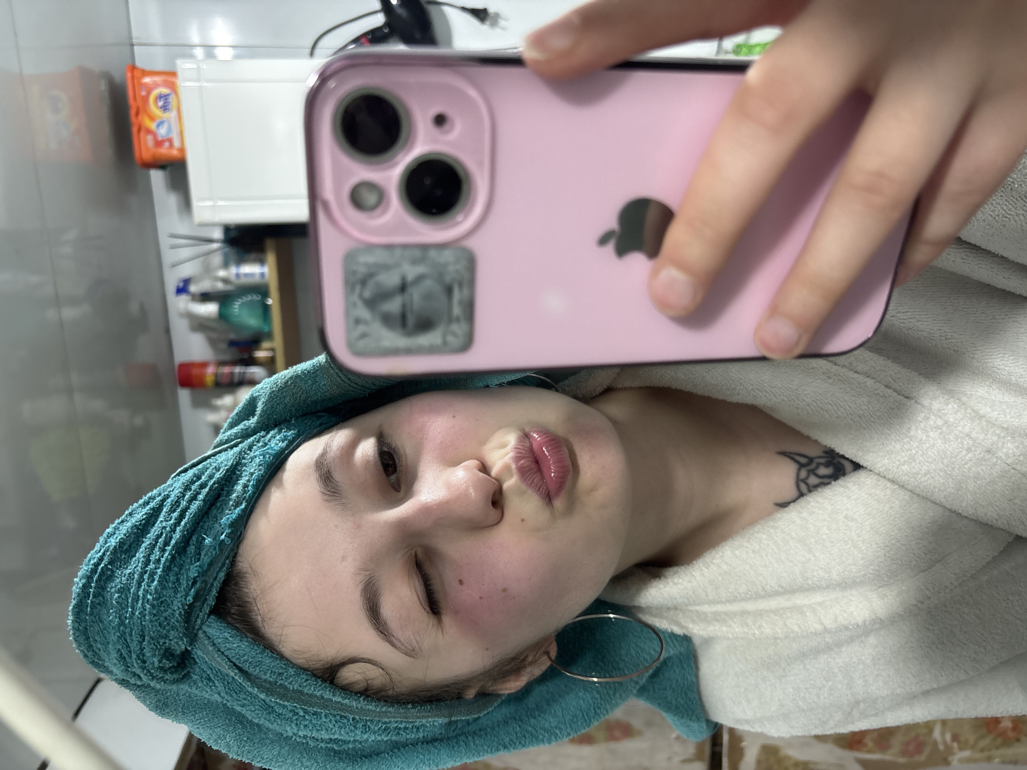 A woman is taking a selfie in what appears to be a bathroom. She is wearing a white robe and a blue towel is wrapped around her head. She is holding a pink smartphone, showing its camera and the Apple logo. The background shows a sink area with toiletries and a package of Tide detergent. The location in the city of Фоки, Russia cannot be ascertained from the image.