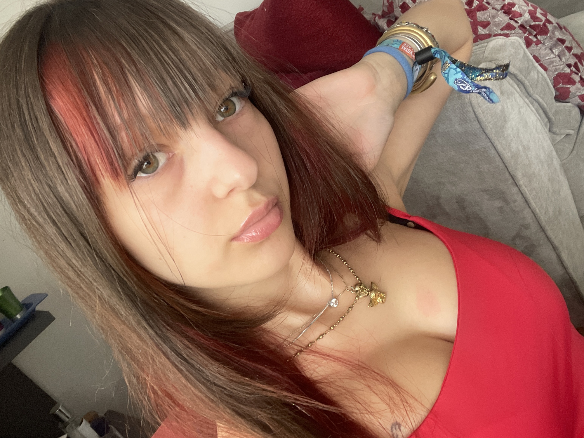 A young woman with long, brown hair with reddish highlights is pictured in a close-up shot. She is wearing a red top and multiple necklaces, one of which features a small golden cherub pendant. Her left arm rests on a grey couch, and she is wearing several bracelets on her wrist, including a blue wristband and a patterned fabric bracelet. The background shows a blurred red pillow and a patterned fabric. There is no discernible location in the city of London, United Kingdom.
