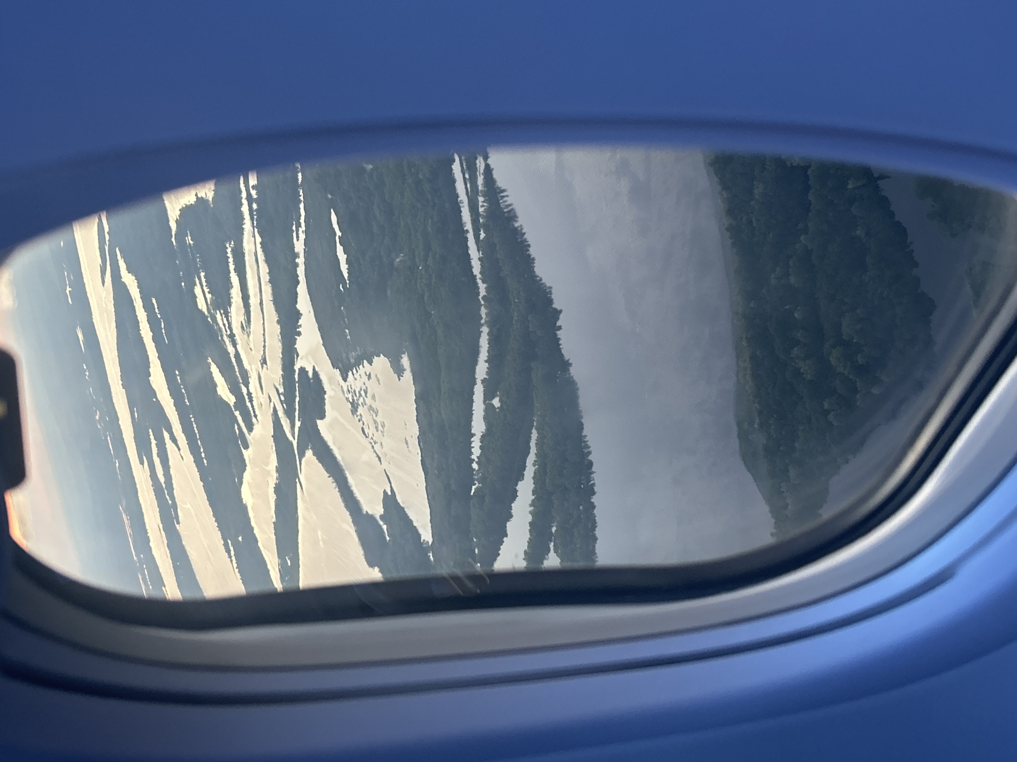 An aerial view through an airplane window shows a vast, sun-drenched river delta near Kokkorevo, Russia. The delta is characterized by numerous islands and channels of water reflecting the sunlight, creating bright, irregular shapes against the darker green of the surrounding forested land. The foreground is dominated by the blue interior of the airplane cabin, framing the landscape. The water appears calm, with the sun casting a warm glow on its surface. The dense green foliage indicates a healthy ecosystem with abundant trees. The overall impression is one of serene natural beauty viewed from a great height.