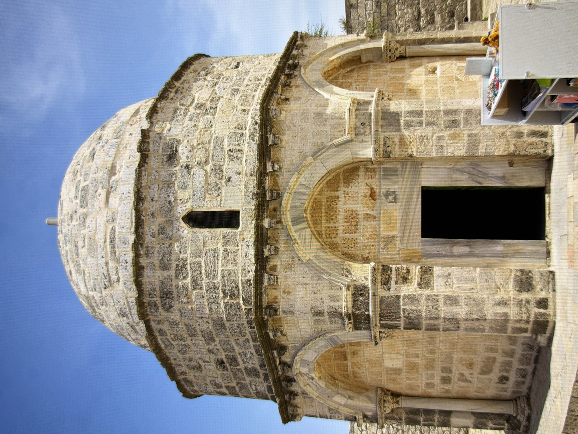 The image shows a circular stone building with a dome, reminiscent of historical architecture. The building is constructed from light-colored stone blocks, with arched doorways and windows. The main entrance is a dark, recessed archway. The dome is also made of stone blocks and is capped with a small finial. In the foreground to the right, there is a white rolling shelf unit displaying various items, including colorful beads and a yellow stuffed animal. The ground around the building is paved with flagstones. The sky is clear and blue. The image is taken on a sunny day. The building does not appear to be located in Kyiv, Ukraine.