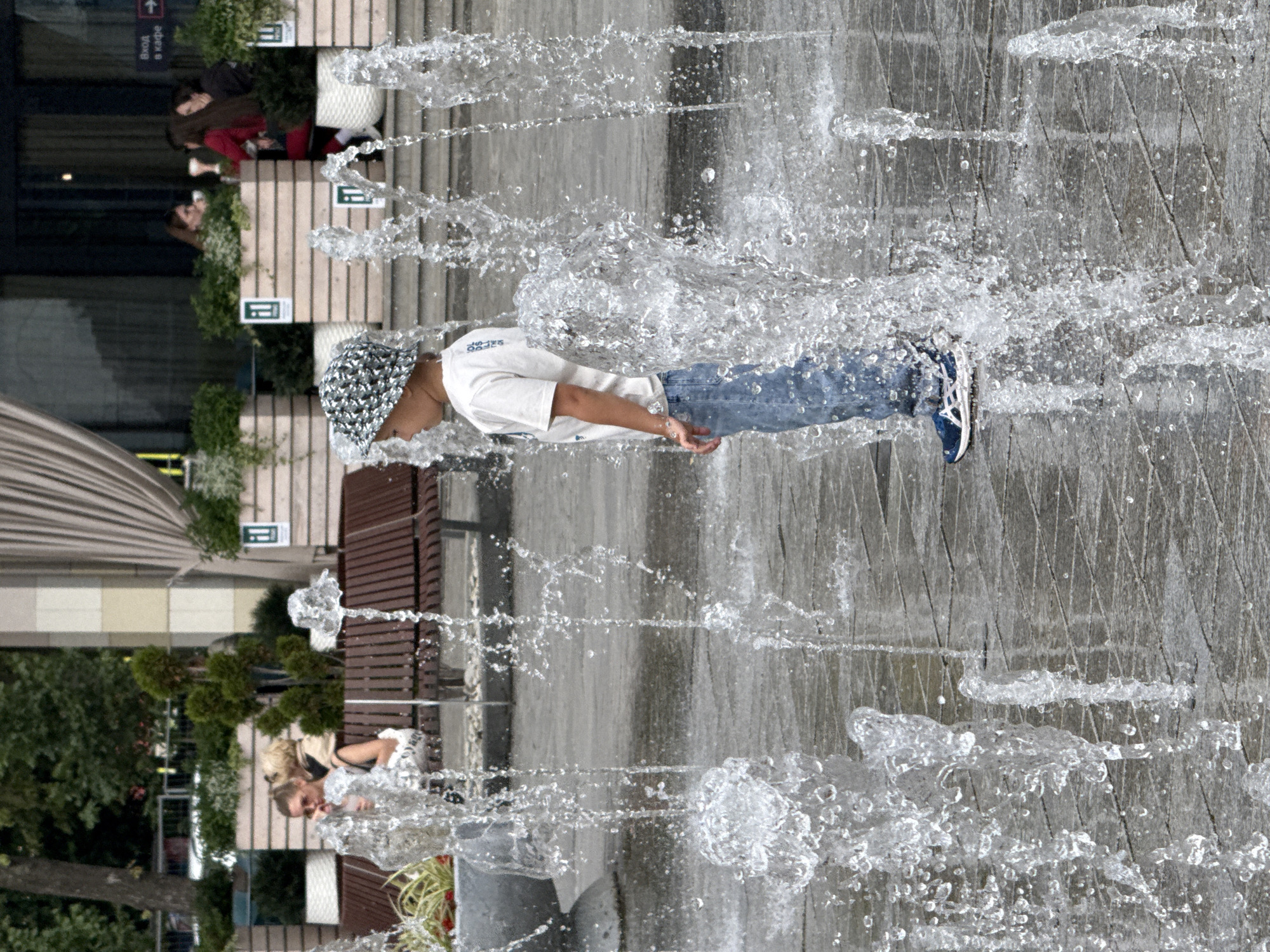 In Moscow, Russia, this media file captures a refreshing outdoor scene with a focus on people interacting with a water feature. The primary subject is a young person wearing a white t-shirt, blue jeans, and a patterned cap, playfully running through a water fountain. The water sprays around them, creating a dynamic and lively atmosphere.
In the background, other individuals can be seen, including a woman with blonde hair sitting and looking towards the water feature, and two people in darker clothing further back. The setting appears to be an urban public space, likely a park or plaza, with paving stones and some greenery visible. The architecture of the surrounding buildings suggests a modern urban environment.
The image is dominated by the spray of water, which creates a blurred effect and adds a sense of movement and exhilaration. The lighting suggests it is daytime, possibly a warm day given the activity. The overall emotion conveyed is one of joy and carefree fun.
Visible text is limited, but on the white t-shirt of the person in the foreground, there is some light-colored text, too indistinct to read. A sign with Russian text, "Вход в кафе" (Entrance to cafe), is visible in the background, providing a specific location clue.
