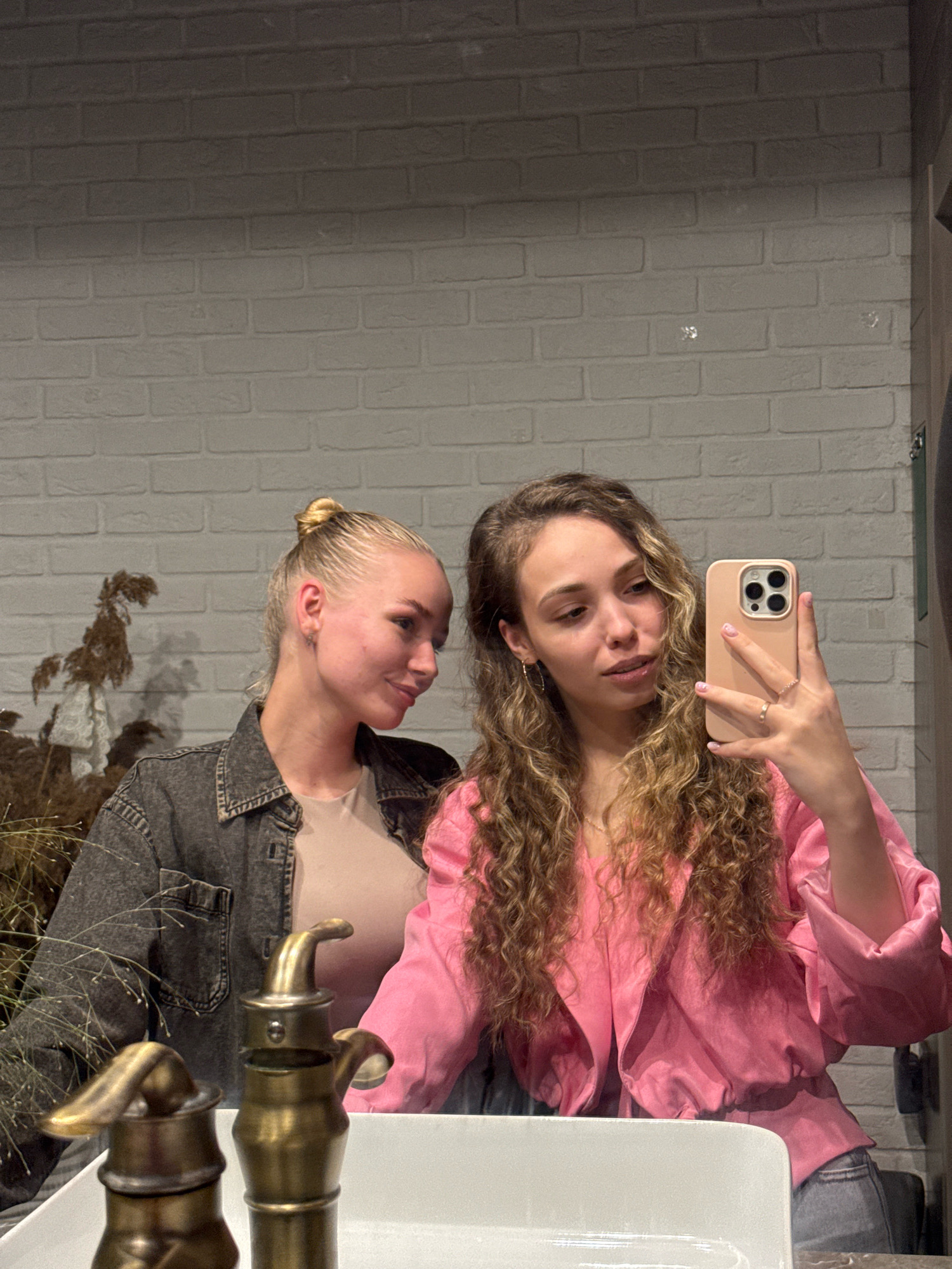 Two women are posing for a selfie in a bathroom with a brick wall. The woman on the left has her hair in a bun and is wearing a denim jacket over a beige top. The woman on the right has curly brown hair and is wearing a pink jacket. She is holding a phone to take the picture. In the foreground, there are two brass faucets in a white sink. The setting appears to be a residential bathroom. There are no specific landmarks that would indicate the location of Chaikovsky, Russia.
