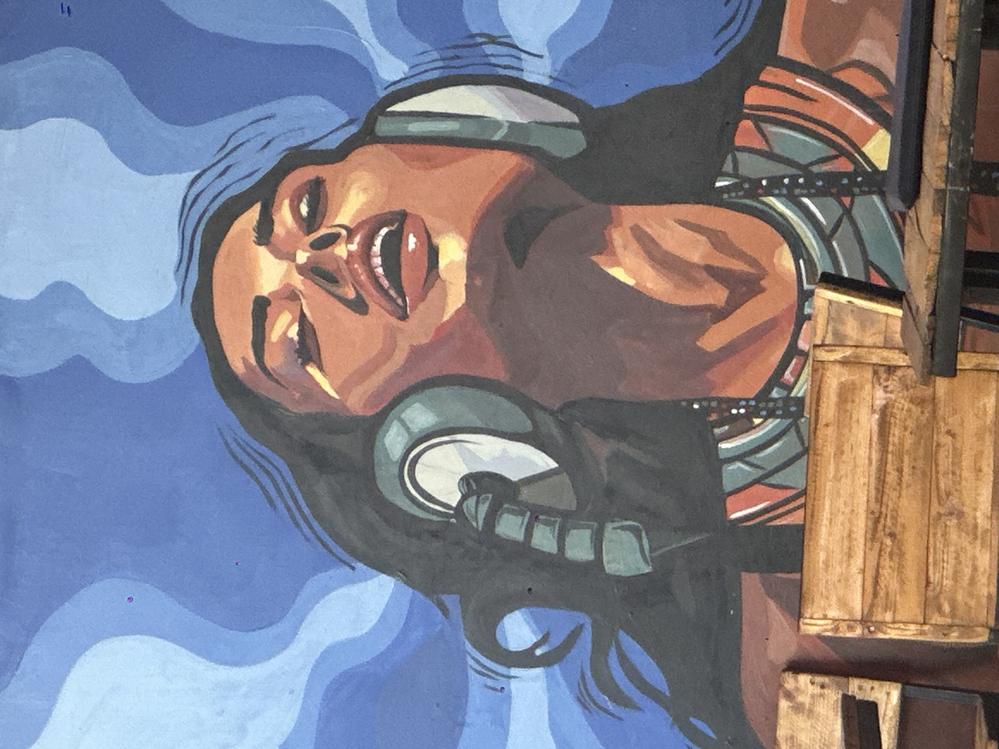The image is a mural painting of a woman wearing headphones, with her head tilted back and eyes closed as if enjoying music. The background is a swirling pattern of blues, evoking a sense of movement or sound waves. The woman's face is painted with warm tones, and her mouth is slightly open. She has long, dark hair that appears to be flowing. Her attire includes a beaded necklace and a patterned top. In the foreground, partially obscuring the lower part of the mural, are wooden chairs and a table. The style of the mural is bold and graphic, with distinct brushstrokes and areas of flat color. There are no visible details that suggest this mural is specifically located in Howayi, Nigeria.