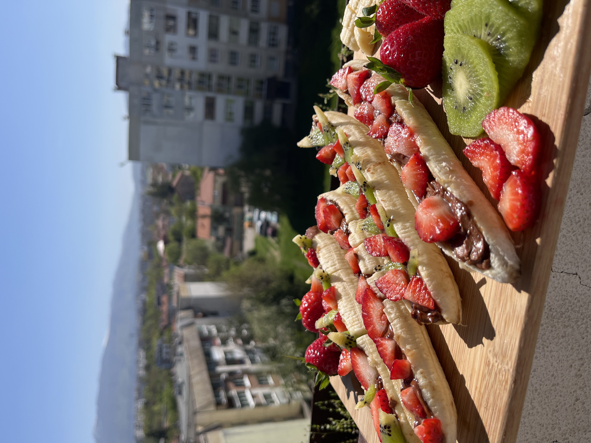 The image displays a series of fresh fruit desserts arranged on a wooden cutting board, set against a blurred backdrop of an urban landscape and distant mountains under a clear blue sky.
In the foreground, several ripe bananas are halved lengthwise and generously filled with a dark chocolate spread, then topped with diced red strawberries and bright green kiwi pieces. These individual servings are lined up neatly on the board. Accompanying them are whole, vibrant red strawberries and several slices of kiwi, showcasing their seeds. The food is sharply in focus and well-lit by direct sunlight, which casts distinct shadows on the cutting board and the light-colored surface beneath it.
The background, largely out of focus, reveals several tall apartment buildings with multiple windows, suggesting an urban or suburban environment. Beyond these structures, a line of green trees separates them from hazy mountains in the far distance. The sky above is a clear, light blue.
The strong sunlight and clear sky indicate a daytime setting, likely morning or early afternoon. The elevated perspective suggests the photograph was taken from a balcony or terrace overlooking the city. No people or visible text are present in the image. The scene suggests a fresh, appealing snack or dessert enjoyed outdoors with a scenic view of Sofia, Bulgaria.