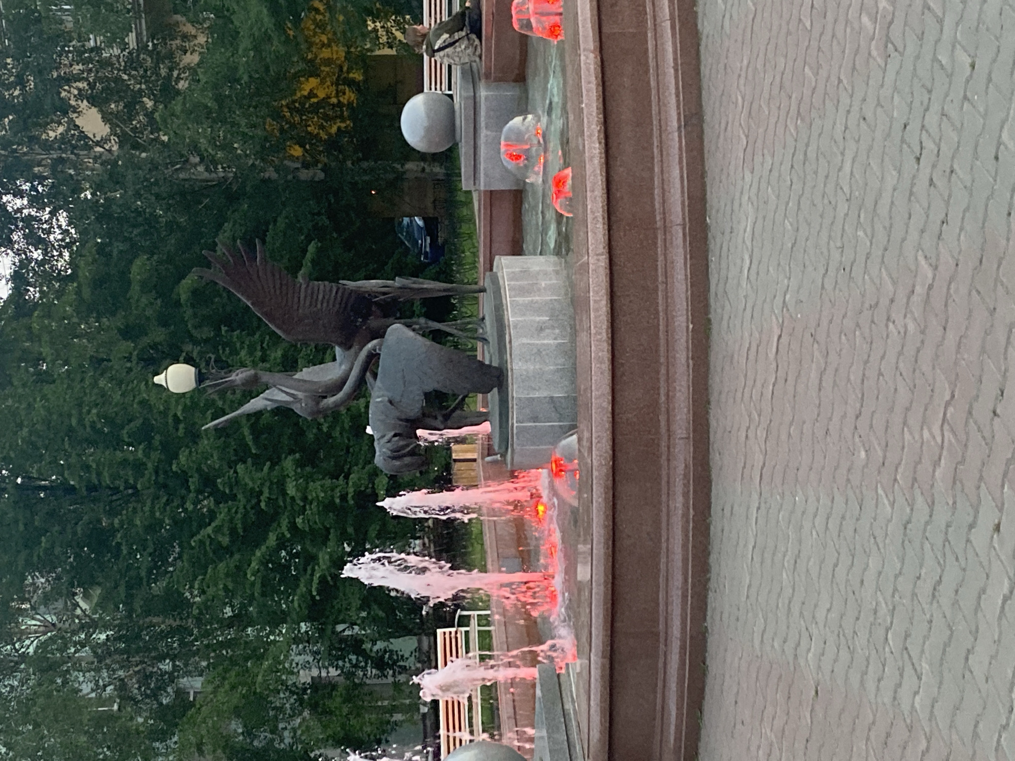 The image shows a fountain with red lights and water jets in front of a bronze sculpture of birds and a human figure. In the background, there are trees, a building, and a parked car. A person is sitting on a bench to the right of the fountain. The scene appears to be in a public park or plaza.