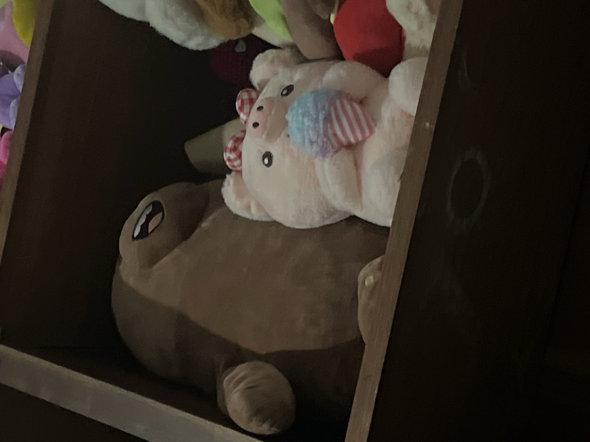 A plush toy of a tan, rounded creature with a wide, open mouth and cartoonish eyes is on the left side of a wooden shelf. To its right, a pink plush pig with red and white checkered bows on its head is holding a plush ice cream cone with blue and pink scoops and pink and white striped cone. The pig has black eyes and a small pink nose. Behind the pig, a plush toy with white fur and a red stuffed object are visible. The shelf is made of dark wood. The image is dimly lit and the background is dark. This image was captured in Santa Ana, Venezuela.