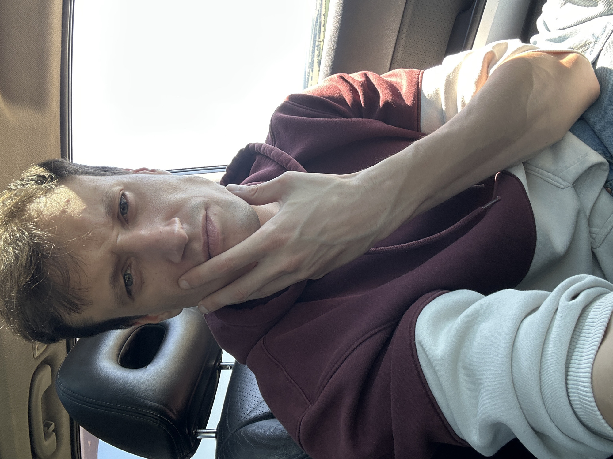 A close-up, eye-level, over-the-shoulder, selfie of a man sitting in the passenger seat of a car. He is wearing a maroon hoodie with white sleeves and has his left hand on his chin, looking directly at the camera. The man has fair skin, short brown hair, and light blue eyes. The interior of the car is visible, including a black headrest and a tan ceiling. The exterior of the car shows a bright, sunny sky and a blurry landscape of fields and trees, consistent with the rural surroundings of Korchivka, Ukraine. The man appears to be in his late 20s or early 30s.