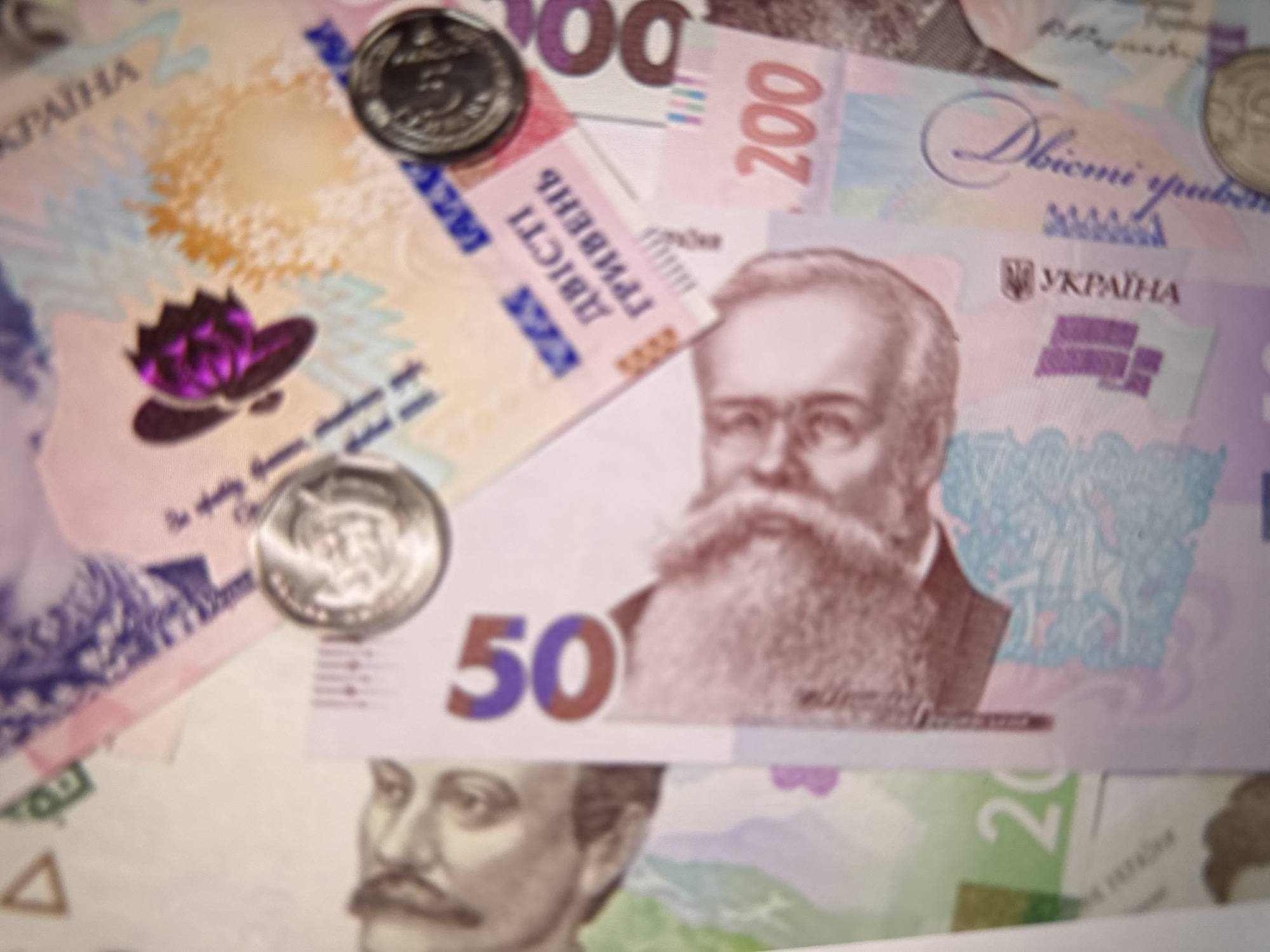 The image displays a collection of Ukrainian Hryvnia banknotes and coins. Prominently featured are banknotes with denominations of 50, 100, and 200. The 50 Hryvnia note shows a portrait of Mykhailo Hrushevsky. The 200 Hryvnia note displays a portrait of Lesya Ukrainka. There are also coins visible, one appearing to be a 5 Hryvnia coin. The background consists of other Ukrainian banknotes in various colors and denominations, some partially obscured. The overall setting is a flat surface, likely a table or counter, and there is no indication of a specific city or location. The image is a close-up shot, focusing on the currency.