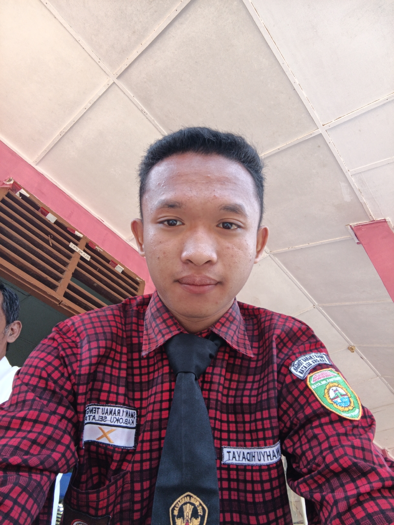 A young man is captured in a low-angle, close-up selfie, his face filling the center of the frame. He wears a red and black plaid shirt with a black tie. A patch on his left breast pocket reads "TAYAQIH UYHAW" in white lettering on a blue background. Another patch on his right sleeve, circular in design, features a shield with an image of a boat on water and a green landscape. A third patch, also on his right sleeve, is a rectangular crest with text that is difficult to decipher. The background reveals a white, paneled ceiling with a pink and red striped structure to the left, resembling part of a building. The ambient lighting suggests an outdoor or well-lit indoor setting. The man appears to be outdoors as suggested by the ceiling structure which is part of a covered area. The presence of school uniforms and a school emblem on the uniform suggests he is a student. The young man is likely in Tanjung Setia, Indonesia.