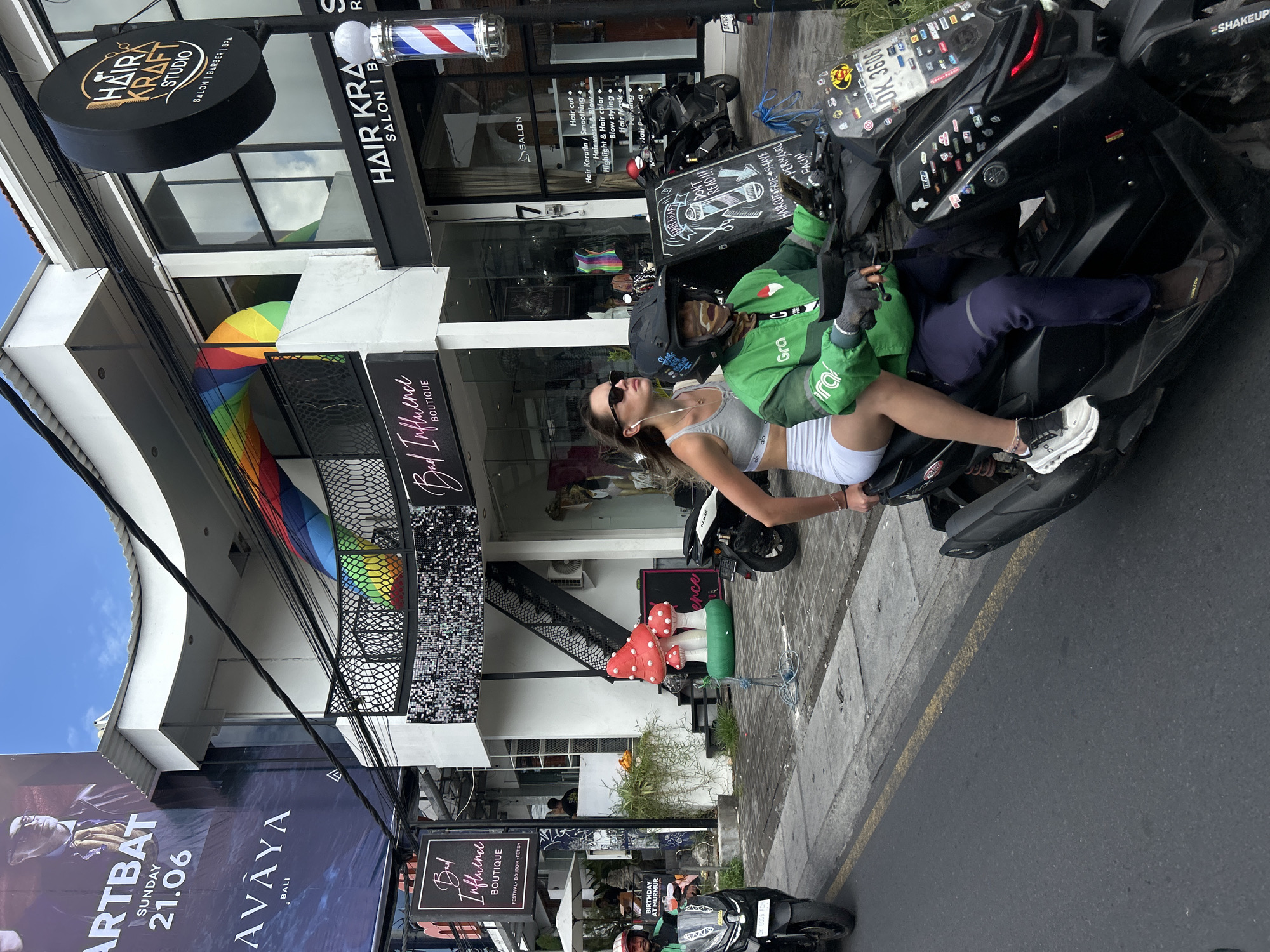 In North Kuta, Indonesia, on a street with shops and businesses, a man wearing a green Grab jacket and dark pants, along with a helmet and face covering, is driving a black scooter. Seated behind him is a woman with sunglasses, wearing a grey sports bra and white shorts, with earbuds in. Both are positioned on the scooter, facing forward on the road. The scooter is adorned with numerous stickers on its front and windshield, and a license plate reading "DK 3606". In the background, buildings with storefronts are visible, including a "Hair Kraft Studio" and a "Bad Influence Boutique". A large poster for "ARTBAT SUNDAY 21.06 AVAYA BALI" is displayed on a building to the left. In front of the "Bad Influence Boutique," a decorative display of red and white mushrooms is present on a green base. The street appears to be paved, with a sidewalk to the right of the road. Other parked scooters are also visible.