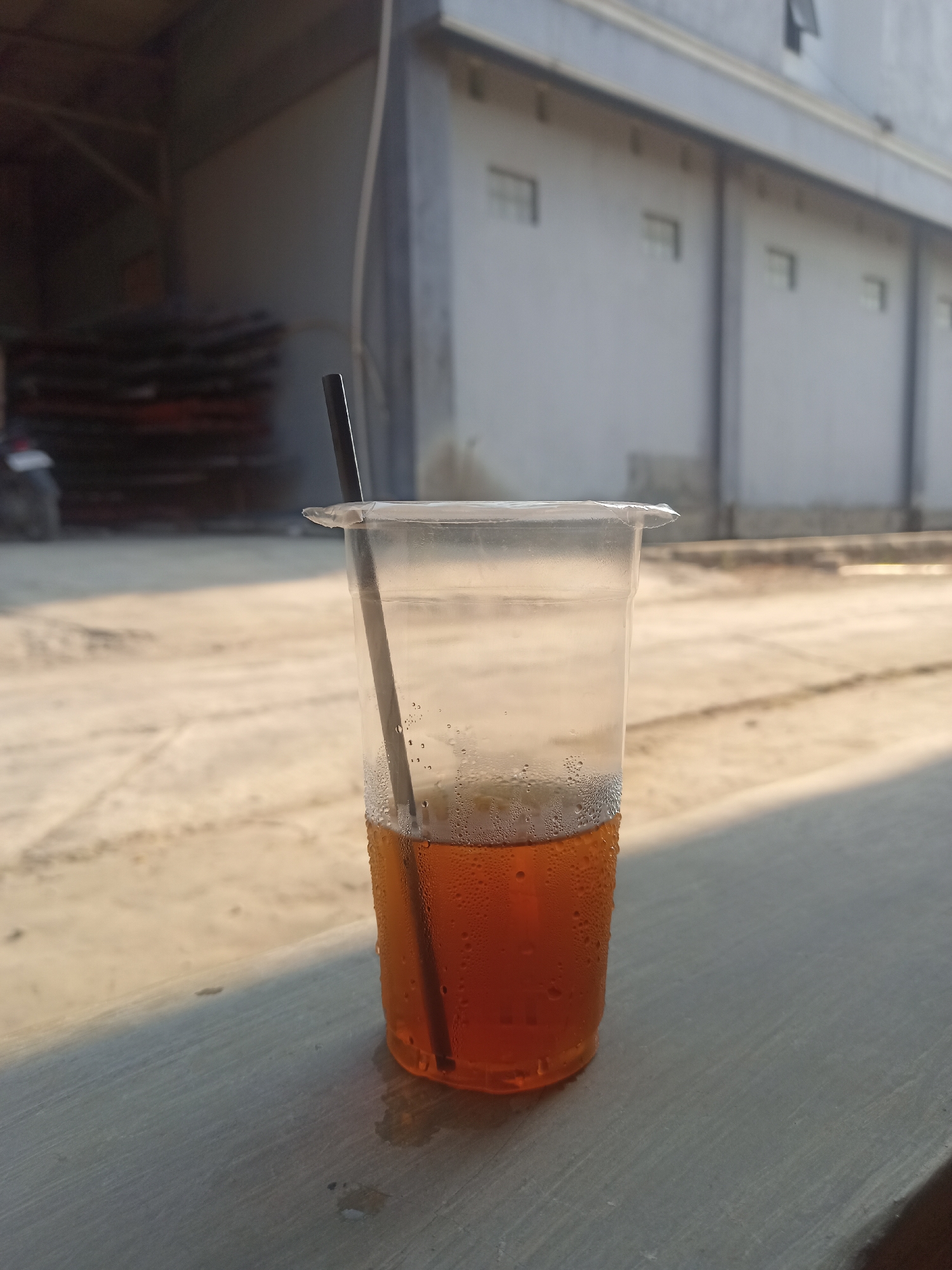 A close-up, eye-level shot shows a clear plastic cup of iced tea with a black straw. The cup is partially filled with a dark amber liquid and condensation is visible on its exterior. The cup is placed on a gray concrete surface in the foreground. Behind the cup, a light brown, sandy area is visible, likely a street or pathway, with shadows cast by sunlight. In the background, a large, light gray building with evenly spaced rectangular windows and vertical support beams suggests an industrial or commercial setting. To the left of the building, a blurred dark area might indicate parked vehicles or stored materials. The overall environment appears to be outdoors in an urban or industrial area, possibly in Karangtalun, Indonesia, given the context.
