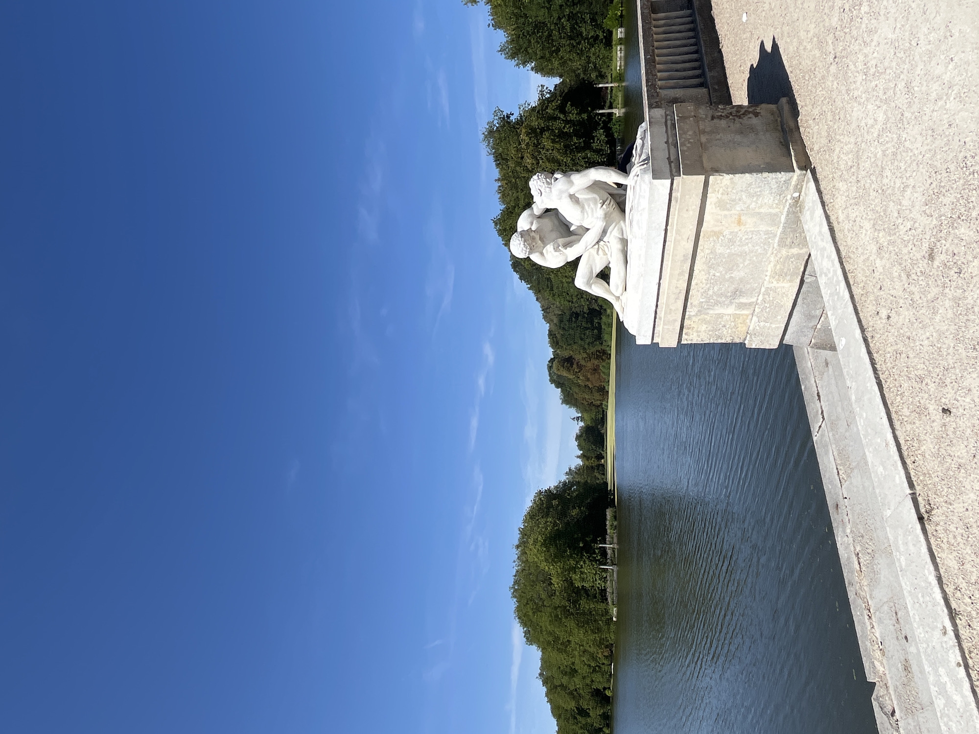 The image depicts a serene outdoor scene with a classical marble sculpture positioned in the foreground. The sculpture, situated on a stone pedestal, features two male figures in a dynamic embrace, rendered in white marble. Behind the sculpture, a tranquil body of water stretches out, bordered by lush green trees and a clear blue sky with scattered wispy clouds. The environment appears to be a park or garden, possibly part of the Château de Rambouillet in Rambouillet, France, given the style of the sculpture and the surrounding landscape. The sunlight casts shadows on the ground, indicating a bright, sunny day.