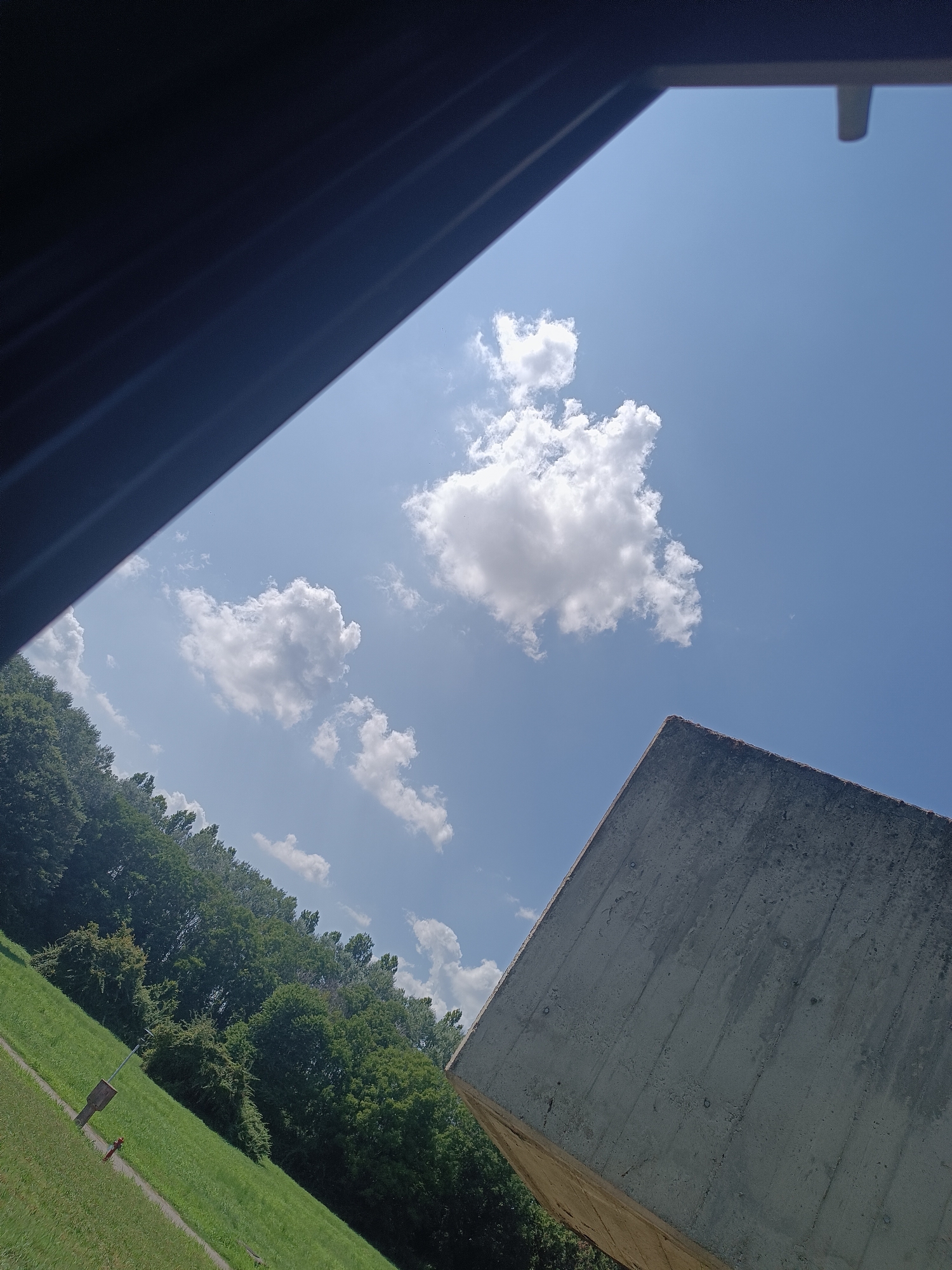 The image shows a view of the sky from a tilted perspective, with a concrete structure and a treeline in the foreground. The sky is blue with scattered white clouds. The concrete structure is a sharp, angular shape, possibly part of a modern building or sculpture, with a rough, textured surface. To the left of the concrete structure, there is a dense line of green trees, which appear to be deciduous. Below the trees, a grassy slope can be seen, with a narrow path winding through it. In the distance, the sky is clear and bright. The overall impression is one of a sunny day, possibly in a park or outdoor recreational area, given the presence of the grass and trees. The perspective suggests the photo was taken from an elevated position, looking upwards.