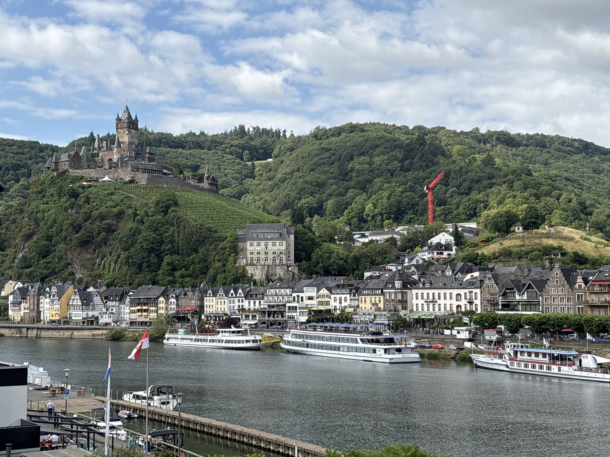 The image shows a panoramic view of the city of Cochem, Germany, situated along the Moselle River. Dominating the left side of the frame is the Reichsburg Castle, perched high on a hill overlooking the city and the river. The castle is a large, medieval structure with multiple towers, battlements, and a reddish-brown tiled roof. Green vineyards cover the slopes leading up to the castle.
Below the castle and stretching along the riverbank is the town of Cochem, characterized by its traditional German architecture. Buildings are predominantly multi-story, with steep, dark roofs, white or light-colored facades, and ornate gables. Many of these buildings appear to house shops, restaurants, or hotels, as indicated by awnings, outdoor seating, and visible signage. Trees line the riverbank, providing a green buffer between the buildings and the water.
The Moselle River occupies the foreground and midground of the image. Several boats are visible on the water. Two large white river cruise ships are moored along the town's promenade. The ship on the right has "KD" written on its side, indicating it belongs to the Köln-Düsseldorfer fleet. Other smaller boats and a wooden pier are also present.
In the background, the landscape is characterized by rolling, heavily forested hills that rise steeply behind the city. A bright red construction crane is visible on the hillside to the right of the castle, suggesting ongoing development or renovation in the area. The sky is partly cloudy, with patches of blue sky interspersed with white, fluffy clouds. Two flags, one with red and white stripes and a crest, and another with blue and white stripes, are visible flying from poles in the foreground. A few people are visible on the pier in the foreground, and some appear to be seated at outdoor tables. The overall scene depicts a picturesque European river town with historical landmarks and vibrant activity.