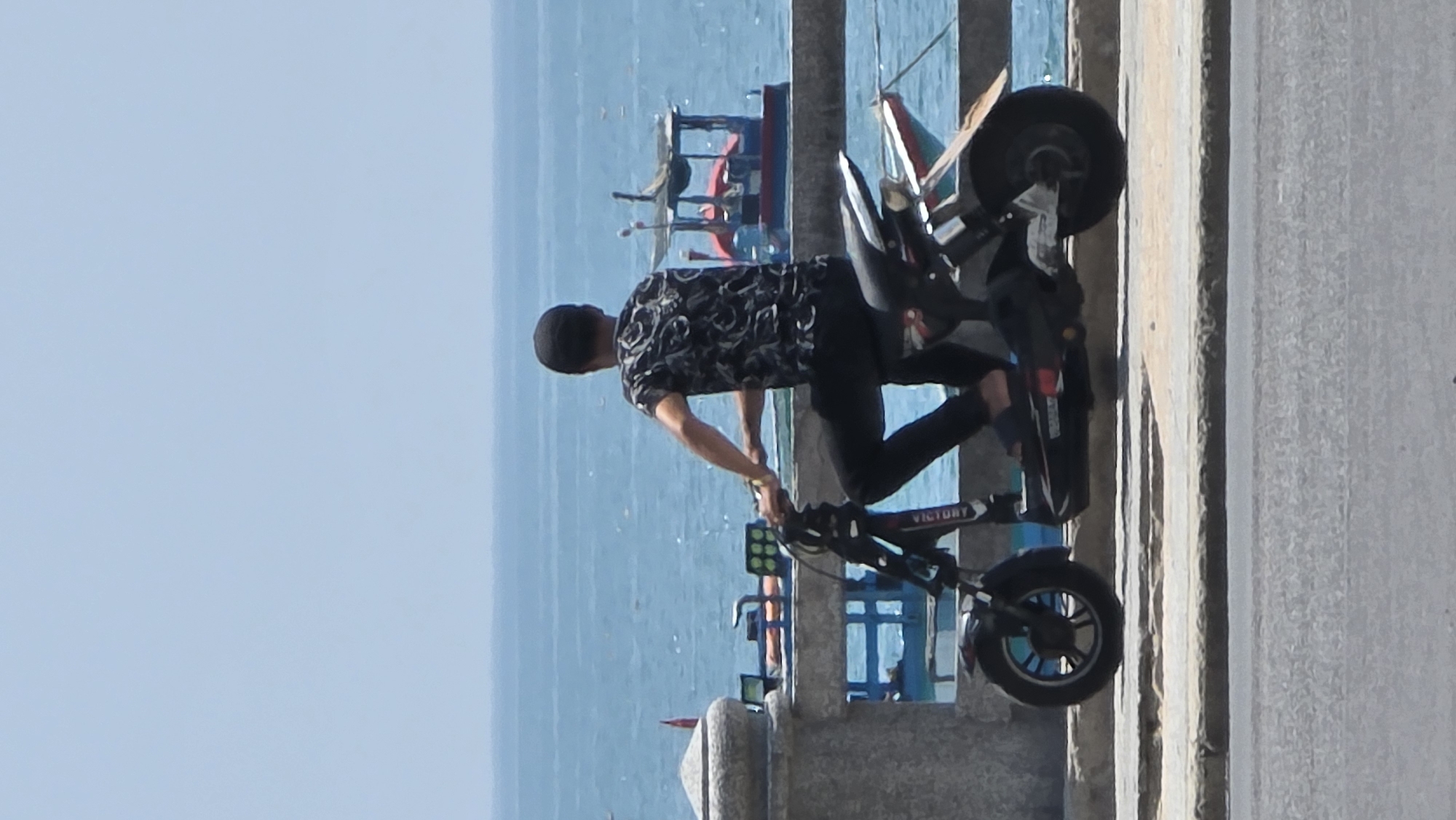 A person is shown standing on a pier in Bắc Nha Trang, Vietnam, next to an electric scooter. The person is wearing a black hat, a dark-patterned shirt, and black pants. The scooter is primarily black, with "Victory" displayed on its frame. In the background, the blue ocean is visible, along with a few boats near the pier's edge. The pier itself is made of concrete. The image is rotated 90 degrees clockwise.