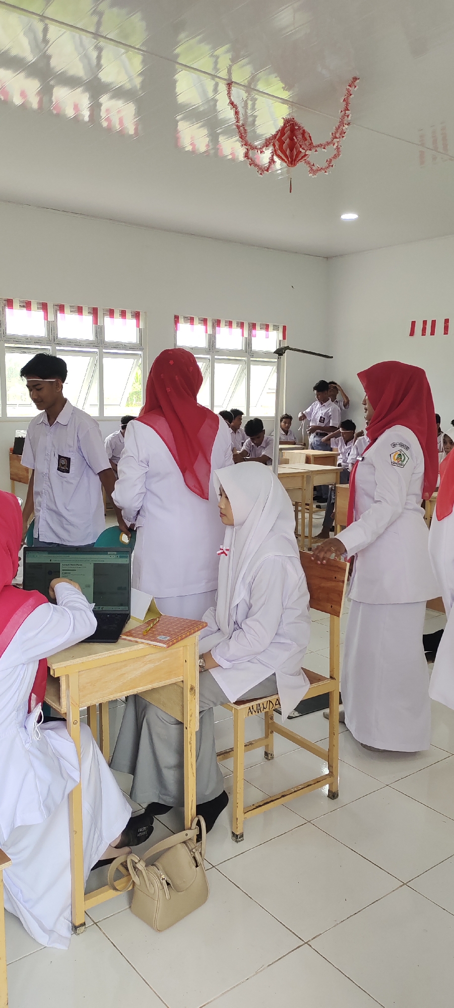 In a classroom in Langnibong, Indonesia, several students wearing white uniforms and headscarves are gathered around desks. A boy in the foreground, wearing a white collared shirt and dark pants, is looking down and to his left. Beside him, a girl in a white headscarf and uniform is seated at a desk, interacting with a laptop. Another student, also in a white uniform and red headscarf, stands behind her, appearing to point at the laptop screen. A third student in a red headscarf and white uniform stands further back, facing away from the camera. The room has white walls and a white tiled floor. Windows with white frames and red and white striped curtains are visible in the background. Red paper decorations are suspended from the ceiling. A beige handbag rests on the floor next to the seated girl's desk.