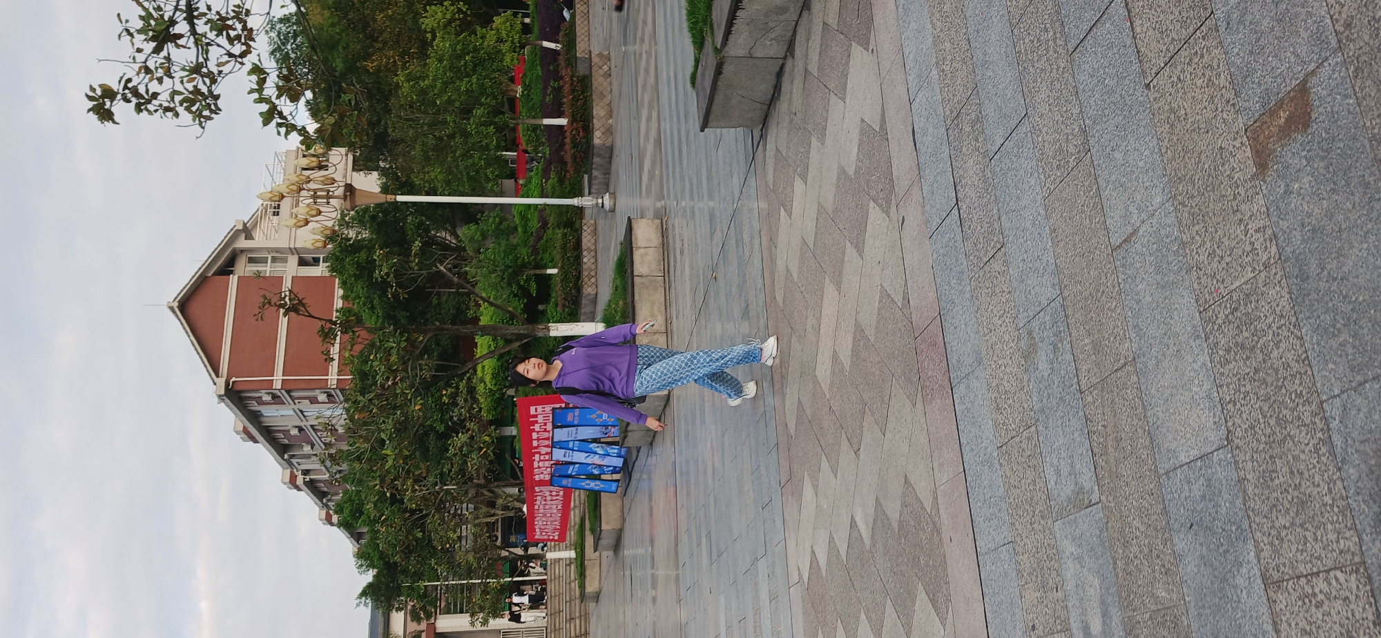 A woman is walking on a paved walkway in Chongqing High-tech Zone, China. She wears a purple sweatshirt and patterned blue pants. Behind her are trees, a building with a red brick facade, a street lamp, and signage. The sky is overcast. The walkway is made of gray and beige pavers laid in a herringbone pattern.