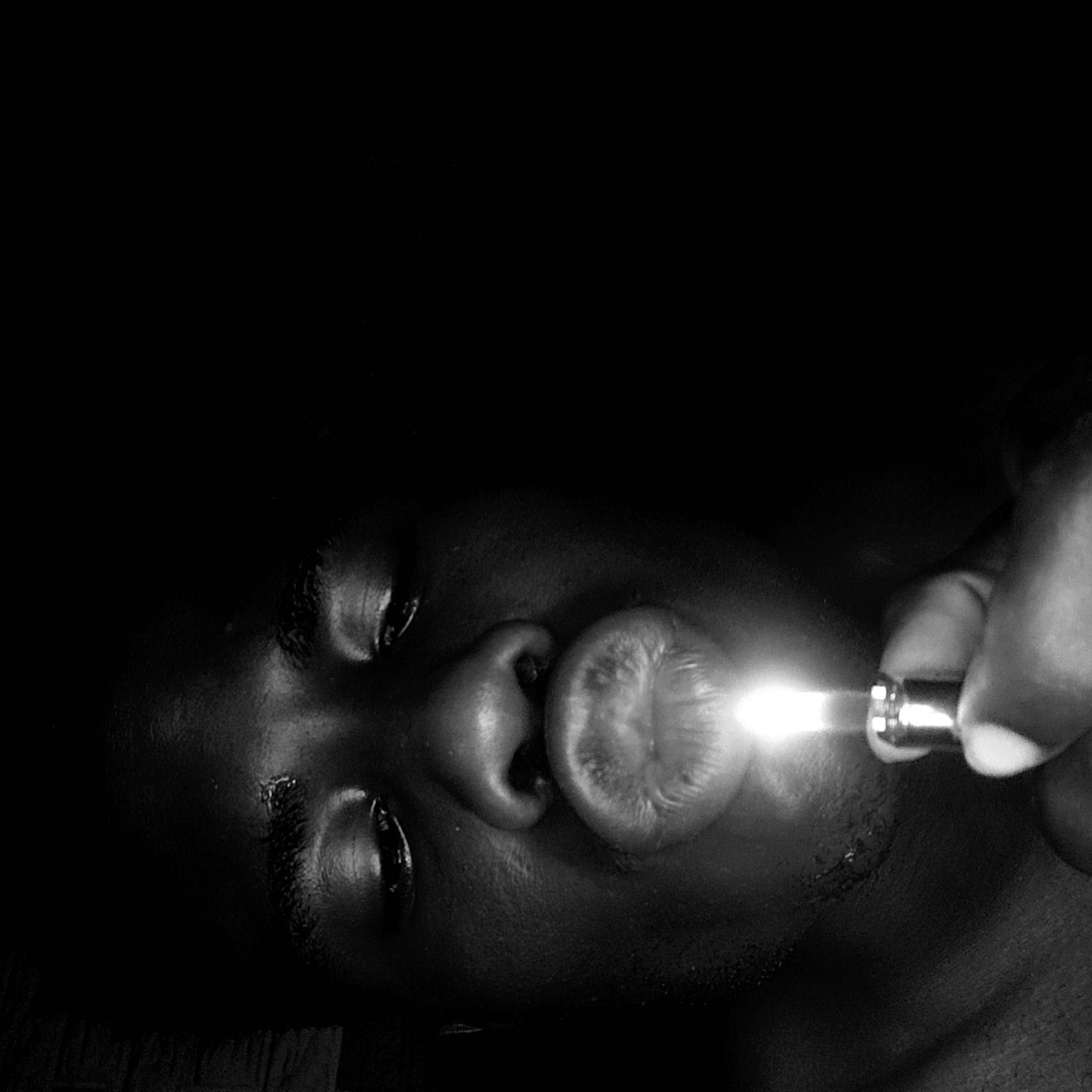 A close-up black and white image shows a person's face from the nose down. The person is dark-skinned. A hand holds a small, cylindrical object with a bright light emanating from the end, pointed towards the person's pursed lips. The background is black and undifferentiated. The location of Narayi, Nigeria is not verifiable from this image.