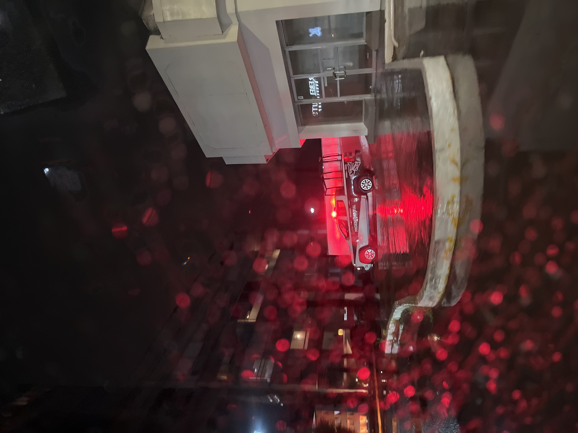 The image shows a fire truck parked in front of a building at night in San Bernardino Tlaxcalancingo, Mexico. The building has glass doors and windows with writing on them. The view is partially obscured by what appears to be raindrops or other red bokeh. Streetlights and other buildings are visible in the distance.