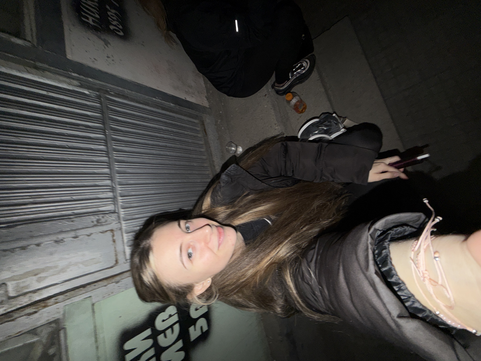A young woman with long, light brown hair is lying on the ground next to a closed metal shutter. She is wearing a dark-colored jacket and facing the camera. Next to her, another person wearing dark clothing is partially visible. Nearby, there is a water bottle and a vape device. Graffiti is visible on the wall next to the shutter. The location is in an urban area in Plovdiv, Bulgaria.