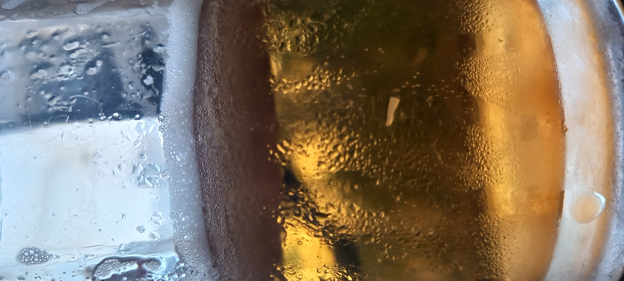 A close-up, vertical photograph captures a transparent glass containing a golden-amber beverage, likely beer, distinguished by a layer of white foam at its surface. The exterior of the glass is covered in numerous small condensation droplets, indicating a cold temperature.
At the top of the image, the clear rim of the glass is visible with scattered, larger water droplets. Below the rim, a dense layer of white foam with varying bubble sizes rests atop the liquid. The foam transitions into the main body of the liquid.
The central and lower portions of the image display the golden-amber liquid. This liquid is largely obscured by the heavy condensation on the glass's outer surface, creating a textured, slightly blurred effect. Reflections, primarily in hues of golden yellow, green, and dark blue/black, are visible through the liquid and condensation, suggesting ambient light sources or background elements. A distinct, elongated, pale white reflection or object appears within the liquid towards the right-center.
Towards the bottom of the glass, the liquid level tapers slightly, and a larger, solitary teardrop-shaped condensation droplet adheres to the exterior. The immediate surface beneath the glass is not clearly discernible, appearing as a blurred, lighter area. No individuals or specific identifying objects are present in the scene.