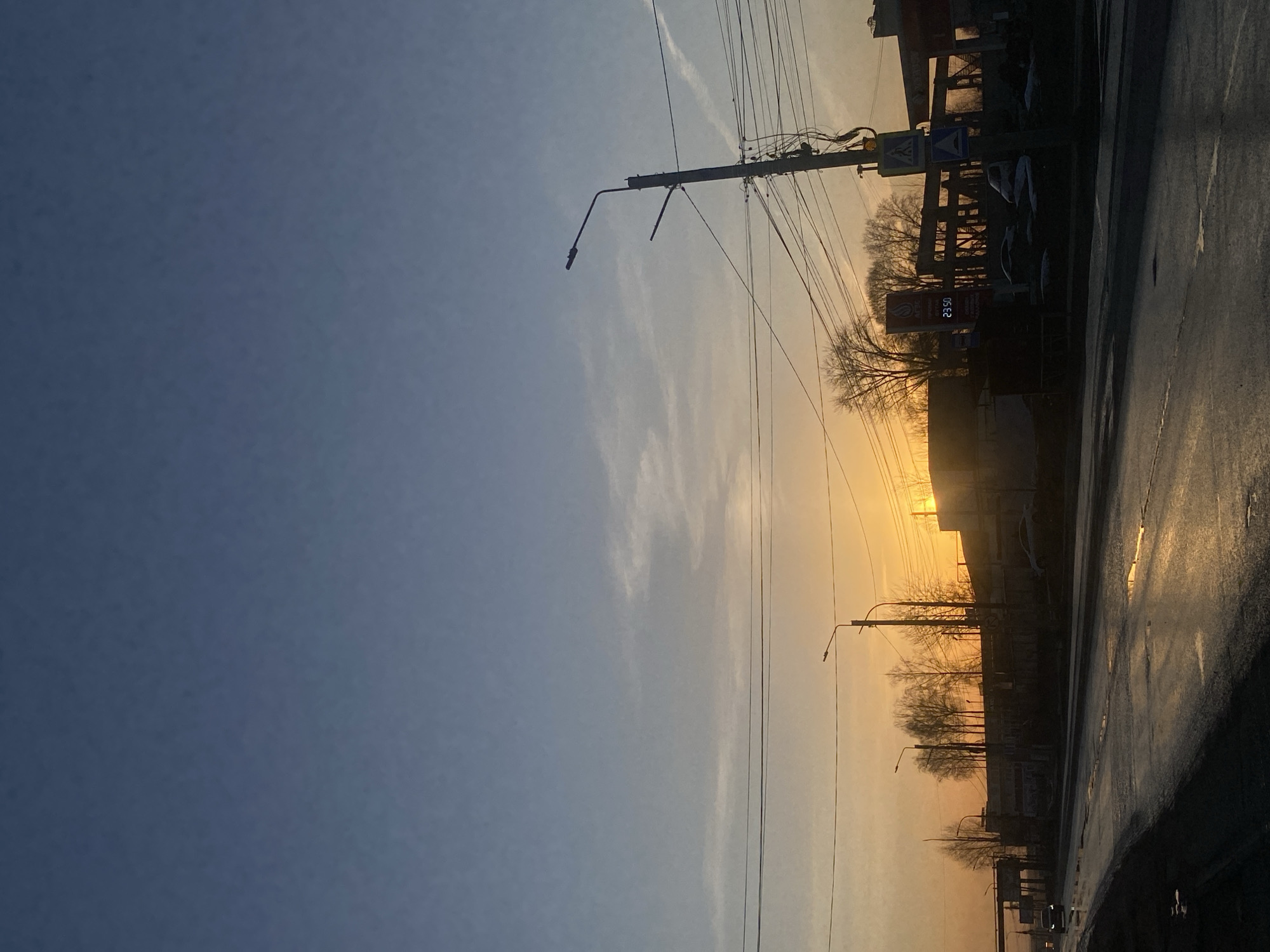 The image captures a street scene in Chaikovsky, Russia, during a sunset. The sky transitions from a clear blue to a warm orange where the sun dips below the horizon. Power lines and streetlights line the street, running parallel to the asphalt, which appears wet and reflective. Buildings and trees are silhouetted against the setting sun. A gas station sign is visible, displaying a price of 23.50.