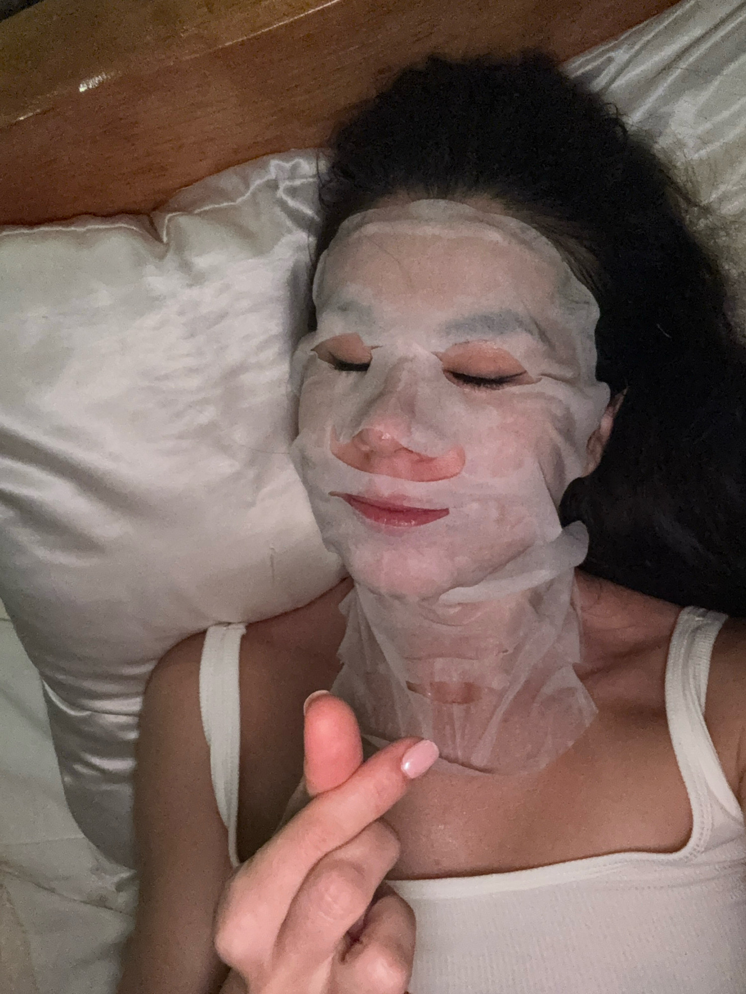 A person with dark hair is lying on a satin pillow with a sheet mask covering their face. They are wearing a white tank top. Their left hand is held up in front of their chest, forming a heart shape with their thumb and index finger. The background appears to be a bedroom. This image was taken in Hiệp Thạnh, Vietnam.