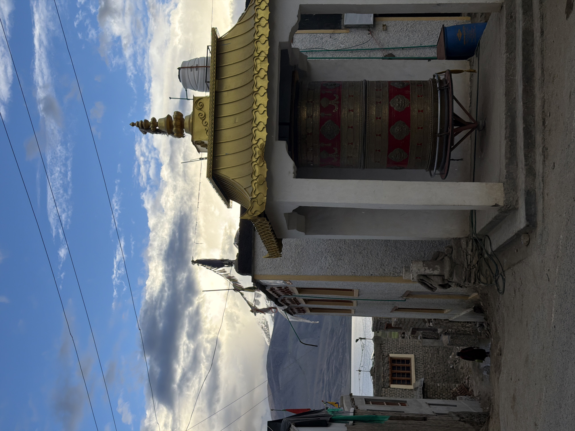In the village of Karzok, India, a prayer wheel with intricate red and gold detailing is positioned in front of a building with a golden roof. The prayer wheel is mounted on a circular stand. To the left of the prayer wheel, a narrow street winds downwards, lined with buildings constructed from stone and plaster. In the distance, a body of water, likely Tso Moriri lake, is visible, bordered by barren, mountainous terrain. A single figure, clad in dark clothing, walks down the street. Overhead, power lines crisscross the sky, which is a mix of blue with scattered clouds and areas of bright sunlight, suggesting either sunrise or sunset.