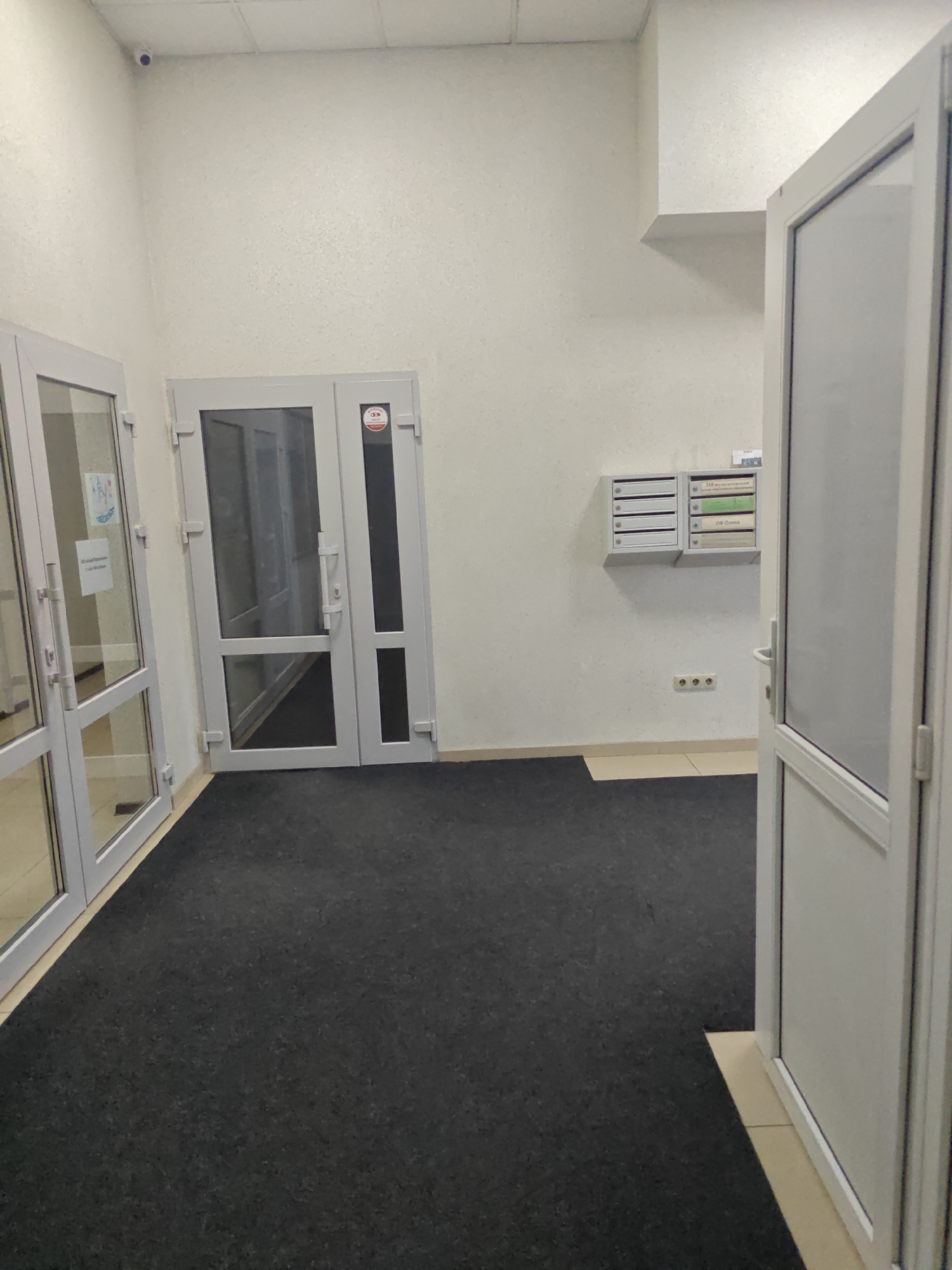 The image shows the interior of a building's hallway or common area, likely a residential building, in Krasnodar, Russia. The scene is captured from a low angle, looking towards two doors and a wall with mailboxes.
On the left, a glass-paneled door with a white frame leads into a space with a checkered floor. A sign with blue and white graphics and Russian text is visible on this door. To the right of this door is another glass-paneled door, also with a white frame, which appears to be open. Through this door, another hallway is visible, also with a checkered floor and a black mat. A red and white circular sticker with an eye graphic is on this door.
The wall to the right of the doors features a set of mailboxes. There are two units, one with five slots and another with three slots, one of which has a green insert and another with a yellow insert labeled in Russian. Below the mailboxes, there are three electrical outlets arranged in a horizontal line.
A black carpet covers the floor in the foreground and extends into the area in front of the doors. The walls are painted white and have a textured finish. A small, white security camera is visible on the ceiling in the upper left corner of the image. The lighting appears to be artificial and from overhead fixtures, contributing to the sterile, functional aesthetic of the space. The overall impression is one of a modern, well-maintained building entrance or corridor.