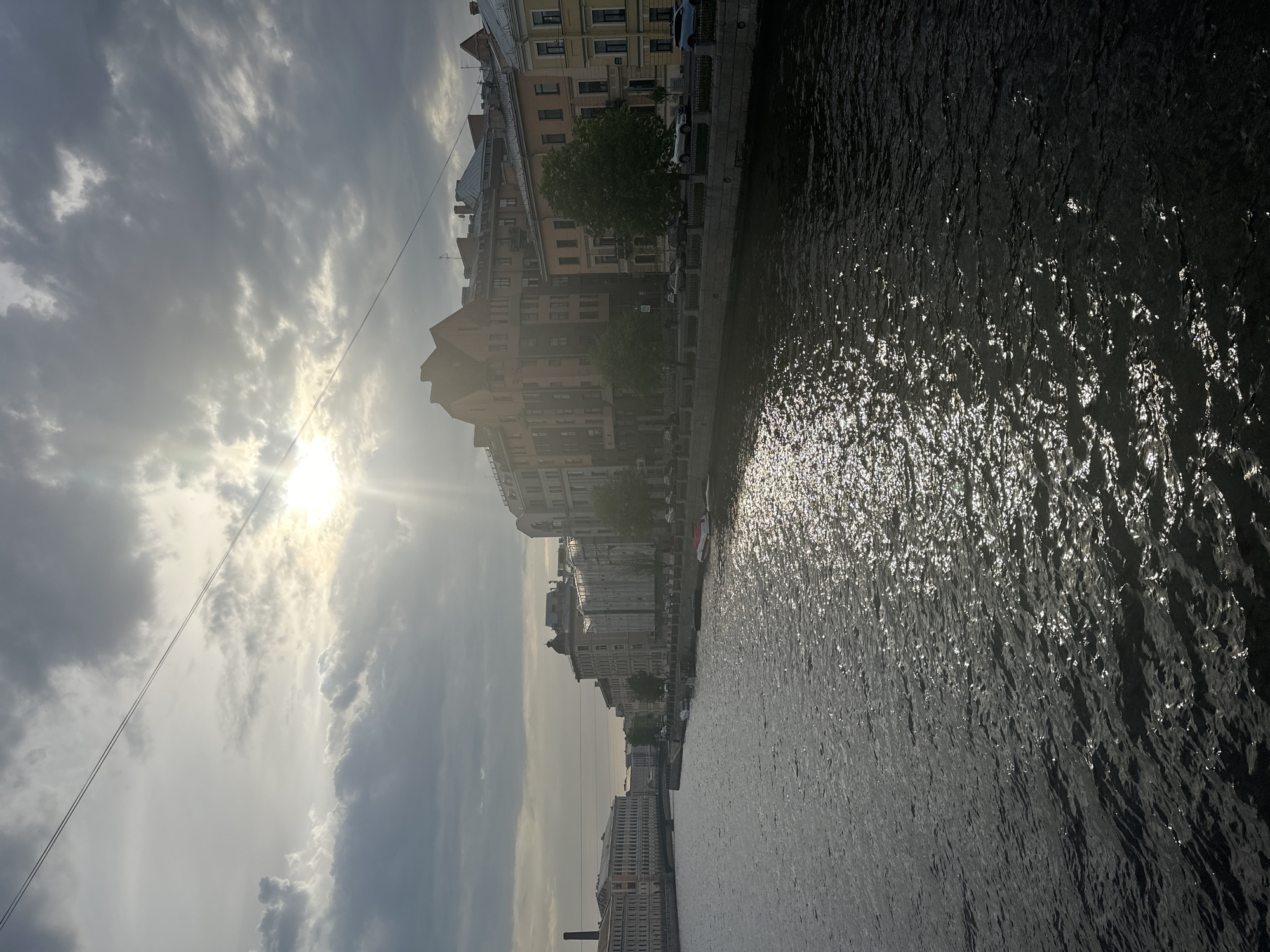 The image depicts a view of Saint Petersburg, Russia, alongside a body of water, possibly a canal or river, with buildings lining the embankment. The water surface reflects sunlight, creating a shimmering effect. Buildings vary in height and architectural style. The sky is overcast, with the sun shining weakly through the clouds. An electrical line is visible against the sky. The photograph appears to have been taken from an elevated position, such as a bridge. There are cars parked along the canal.