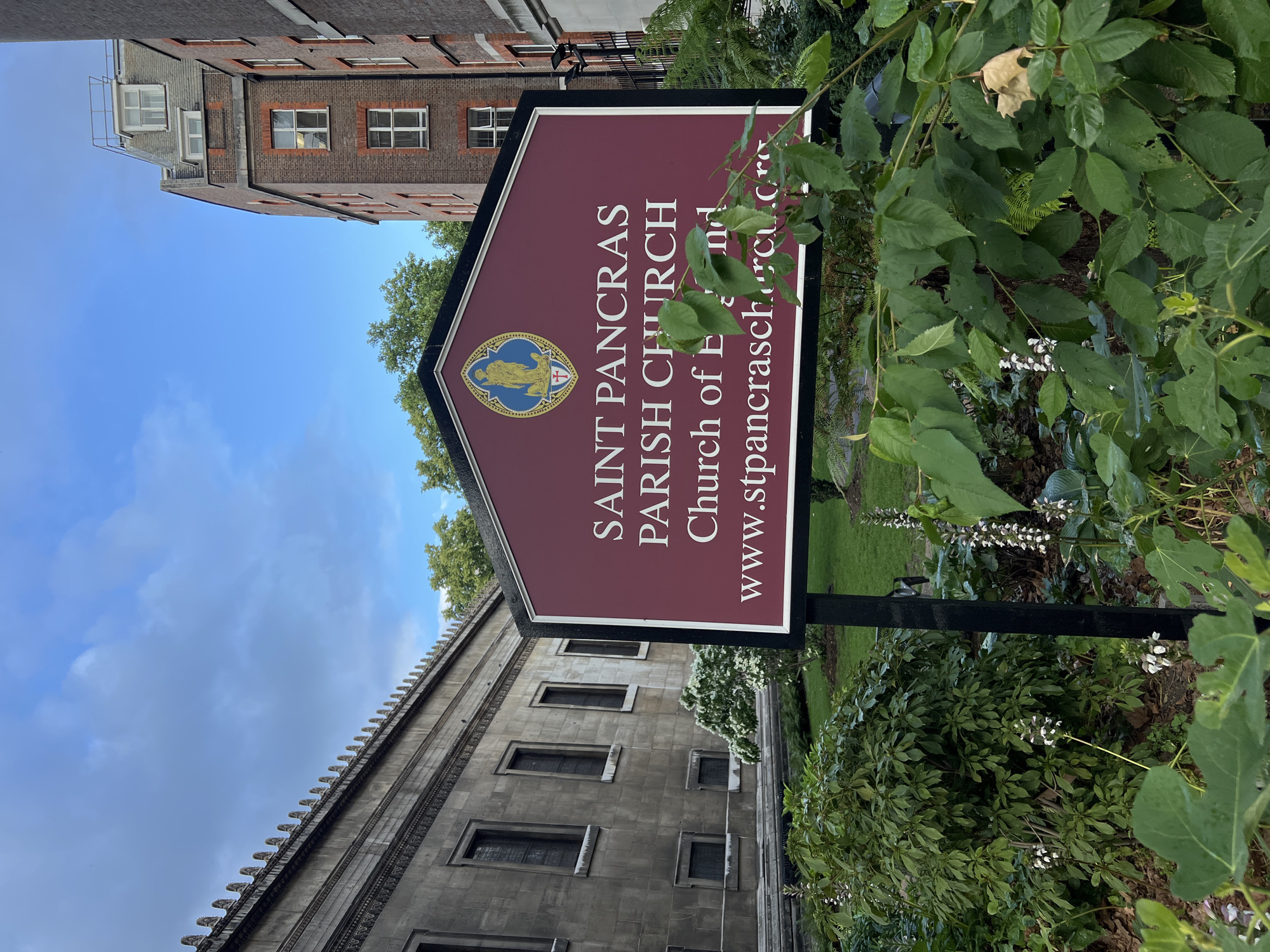 In the foreground, a sign with a maroon background and white lettering reads "SAINT PANCRAS PARISH CHURCH Church of England www.stpancraschurch.org". The sign is supported by black metal posts and is surrounded by green foliage. Behind the sign, a large, ornate stone building with tall, narrow windows stands on the left. To the right of this building, a brick building with multiple windows and a mansard roof is visible. The sky above is a cloudy blue. The scene appears to be in a churchyard or a public space adjacent to a church in an urban environment.
