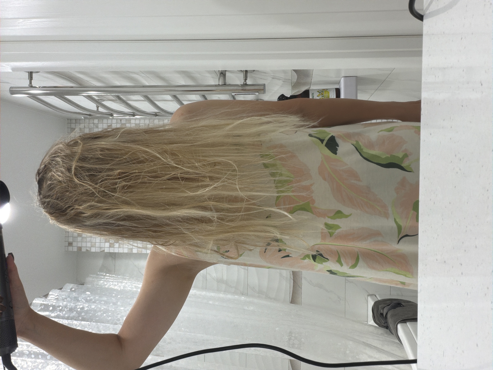 A person with blonde hair is standing in a bathroom and drying their hair with a hair dryer. The person is wearing a floral patterned dress. A shower curtain with a floral pattern is visible behind them. A towel is hanging on the side of the bathtub. A bottle of liquid is on a shelf in the background. The bathroom appears to be clean and modern, with tiled walls and a heated towel rack. The overall scene suggests a routine grooming activity in a domestic setting.