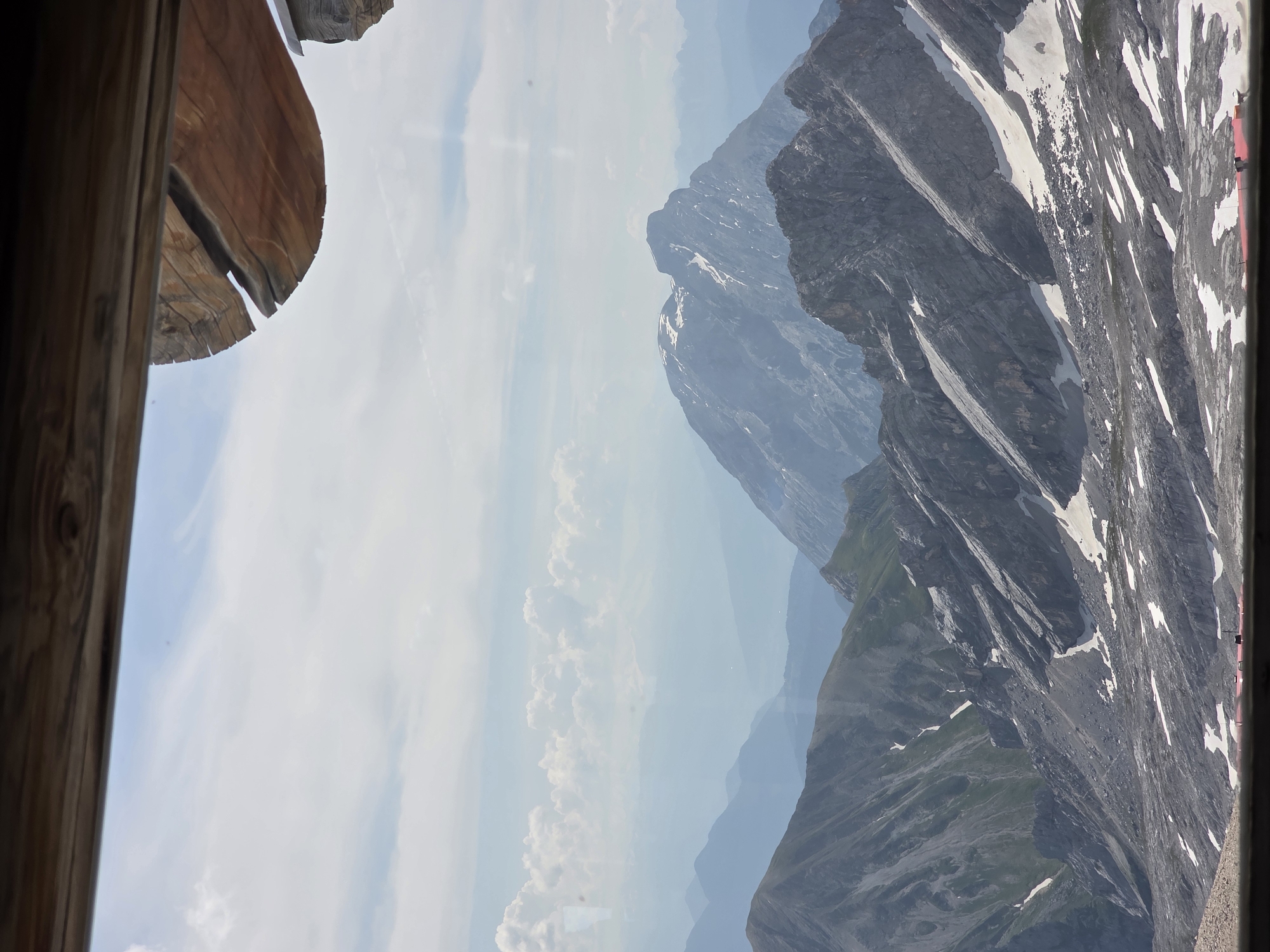 The image shows a panoramic view of a mountainous landscape, likely from a high vantage point. The foreground is framed by a wooden structure, suggesting a balcony, observation deck, or building with a window. The sky is partly cloudy with a light blue hue.
In the middle ground and background, a series of mountain ranges recede into the distance. The closest mountains are rugged and rocky, with patches of snow clinging to their slopes. Further mountains appear softer and more hazy, forming layers of blue and grey due to atmospheric perspective. One prominent peak in the mid-distance has a snow-capped summit.
At the very bottom of the image, a sliver of what appears to be a red surface is visible, possibly a building roof or some man-made structure, with a small dark object in front of it, which could be a vehicle or a person. The overall impression is one of a vast, natural mountain environment, typical of alpine regions. Garmisch-Partenkirchen, Germany, is known for its stunning alpine scenery, including the Zugspitze, Germany's highest peak, which features similar rocky terrain and snowfields. This view is consistent with the mountainous terrain found in and around this Bavarian town.