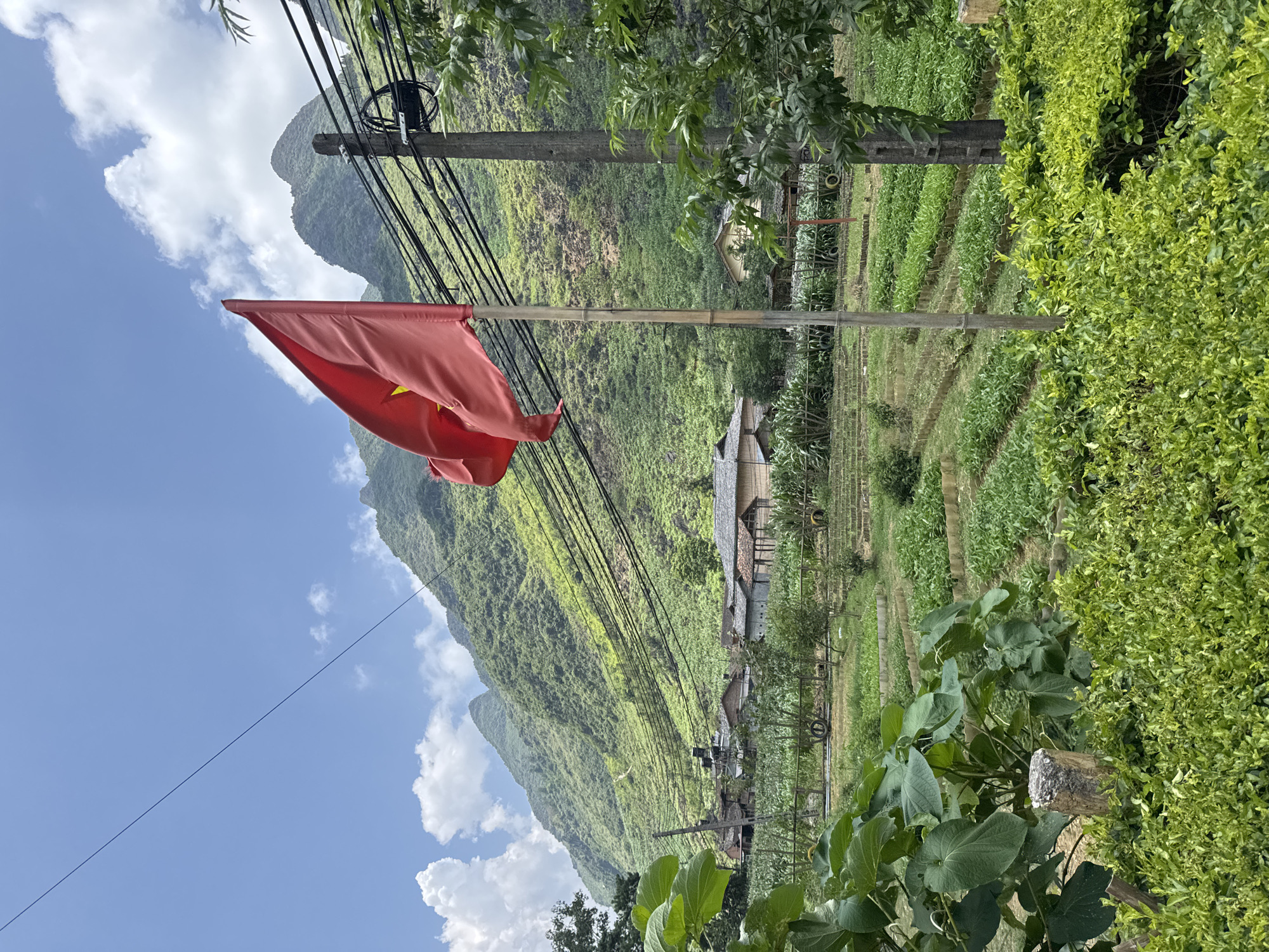 A Vietnamese flag flies near an electric pole in Mèo Vạc Commune, Vietnam. Lush green mountain slopes rise in the background under a blue, partly cloudy sky. Houses with thatched roofs are nestled in the valley amidst terraced fields. Green foliage is visible in the foreground.