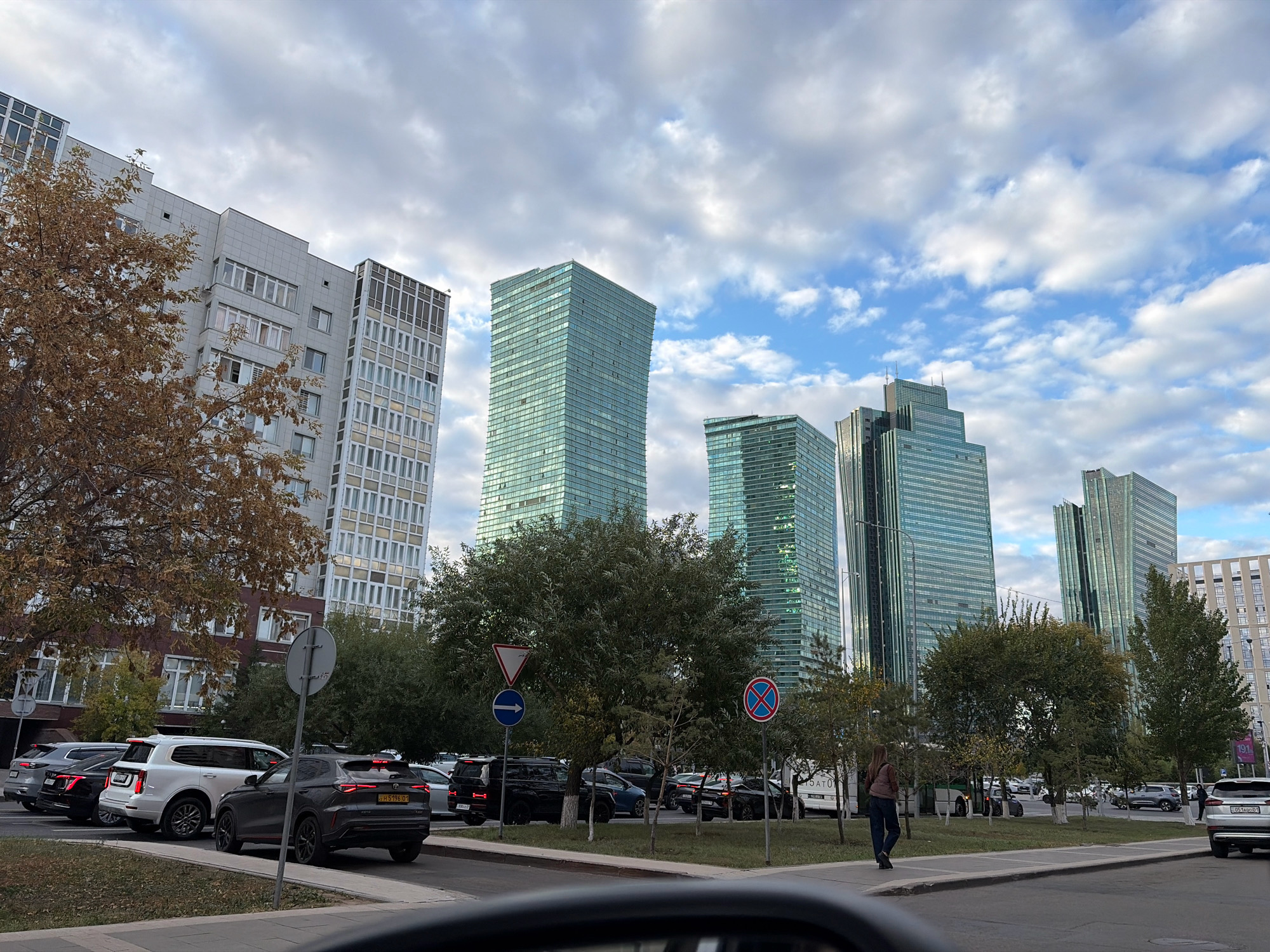 The photograph captures a street scene in Astana, Kazakhstan, likely in the vicinity of the futuristic skyscrapers that characterize the city's modern architecture. Dominating the background are several tall, glass-fronted buildings, including a striking teal skyscraper that tapers towards its peak, and other similar structures. In the foreground, a line of cars is parked along a street, including a white SUV, a dark gray SUV, and a black SUV. A person is seen walking on the sidewalk in the middle ground, facing away from the camera. Trees with autumn foliage are scattered throughout the scene, with some bare branches indicating the season. Traffic signs, including a blue circular sign with a white arrow and a red circular sign with a red bar, are visible. The sky is partially cloudy with patches of blue. The overall impression is that of a bustling urban environment with a blend of modern architecture and natural elements.