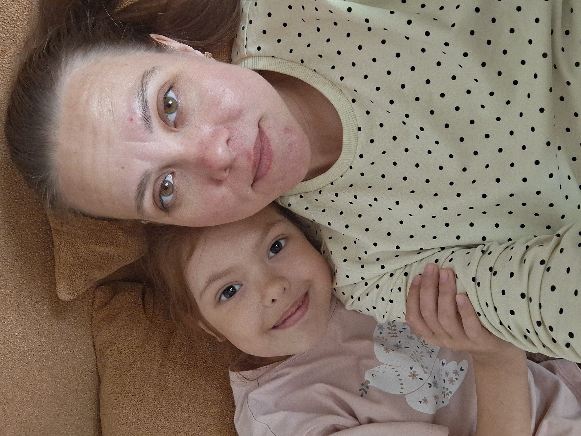 A woman and a young girl are lying close to each other on a brown, textured surface. The woman, positioned above the girl, is wearing a cream-colored top with black polka dots. She has brown hair pulled back and visible freckles on her face. The girl, lying beneath the woman, is wearing a pink shirt with a bird graphic on it. Both are looking directly at the camera with slight smiles. The location is Chaikovsky, Russia.